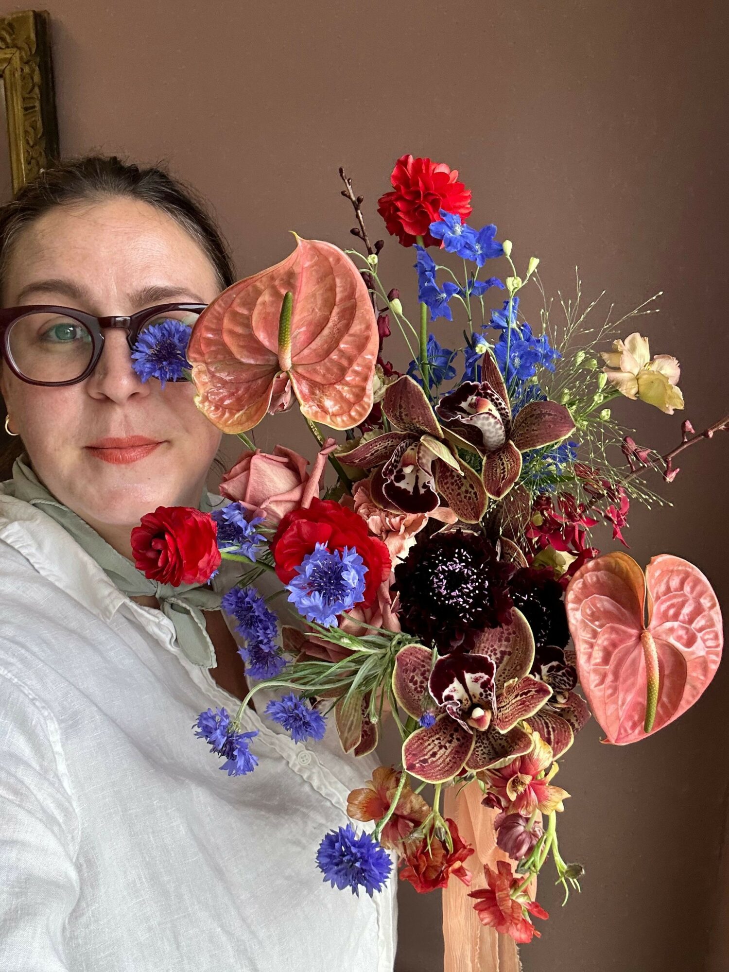 Person with glasses holding a large colorful flower arrangement, partially covering their face, against a plain background.