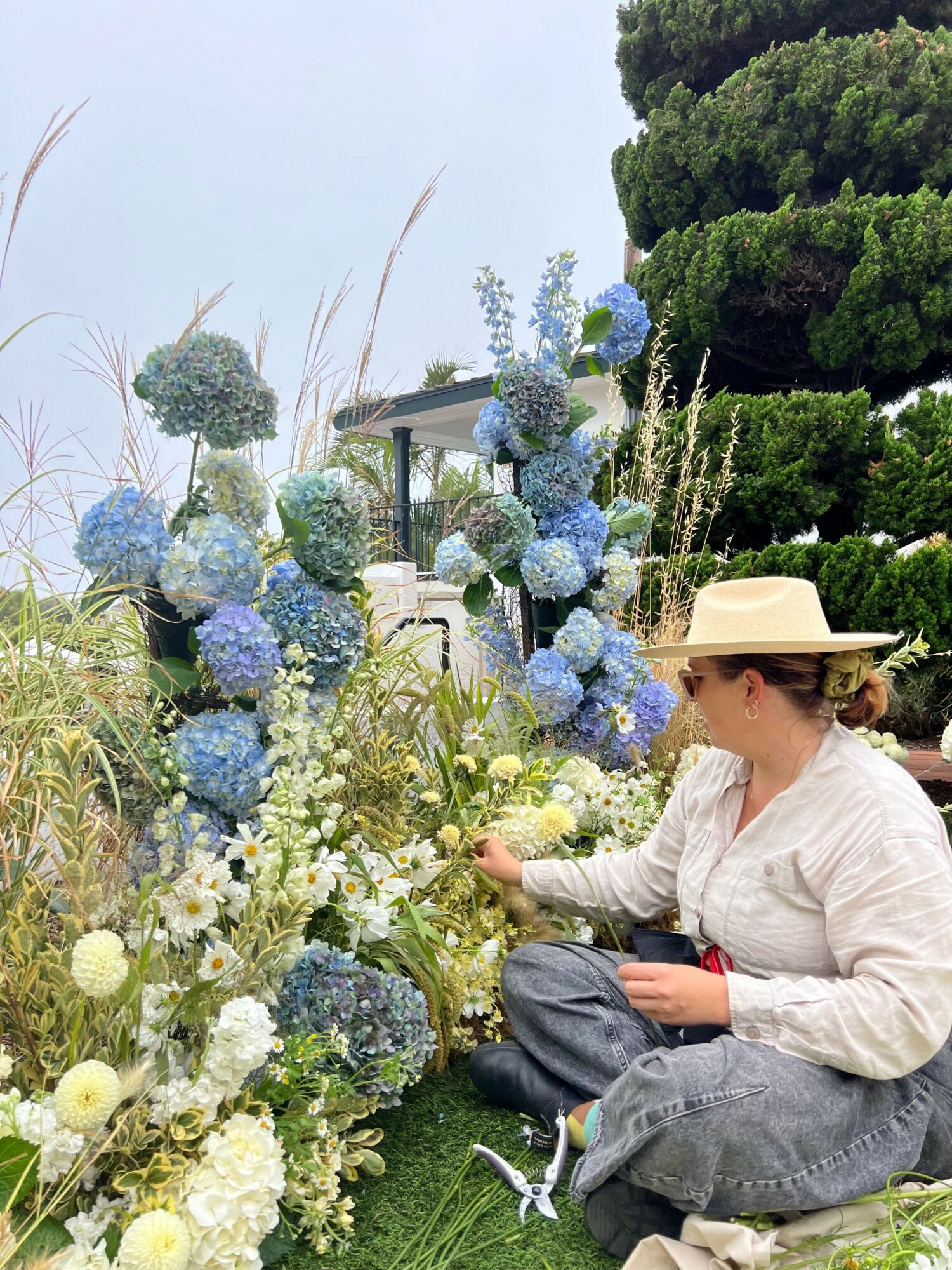Person in white shirt and hat arranging flowers at a memorial site with blue and white floral arrangements.