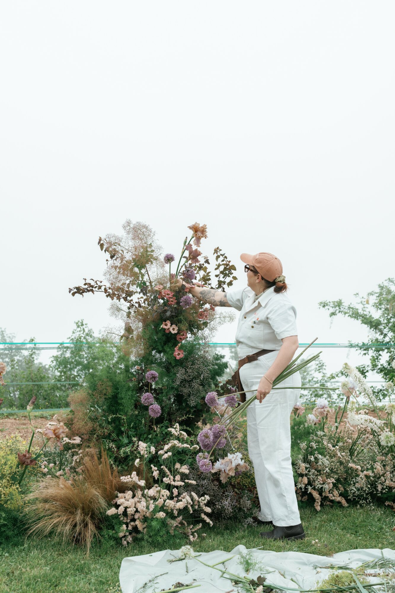 Person in white outfit and cap standing next to a large flower arrangement outdoors, with plants and sky in background.