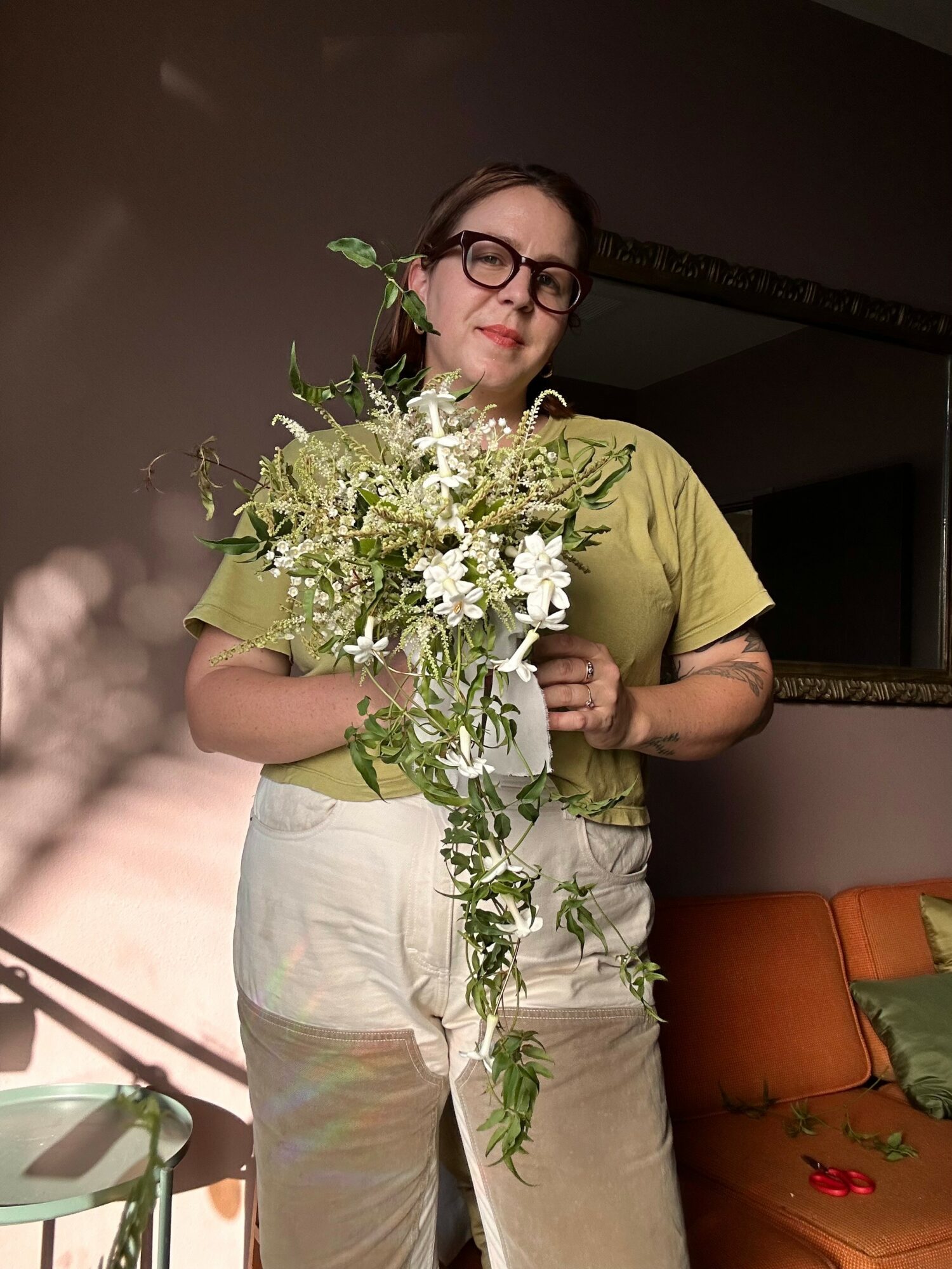 Woman with glasses holding a large bouquet of white and green flowers, wearing a green top and light-colored pants, indoors.