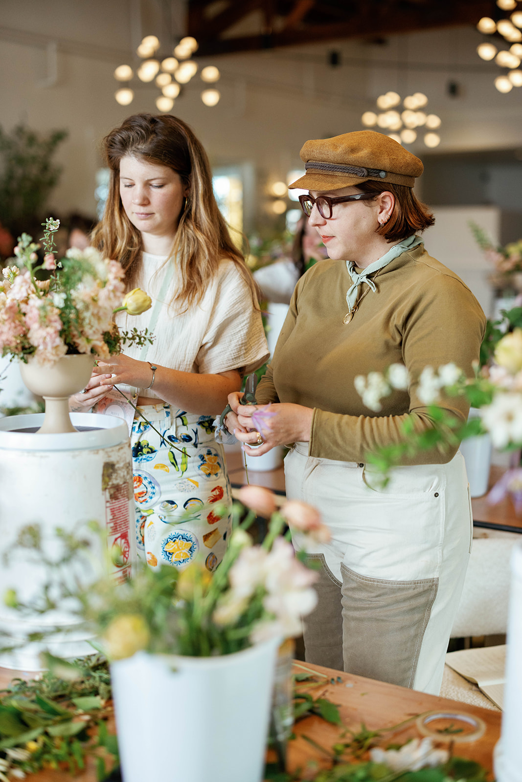 Two women look at flowers in a bright indoor space with hanging lights, one wearing glasses and a hat.