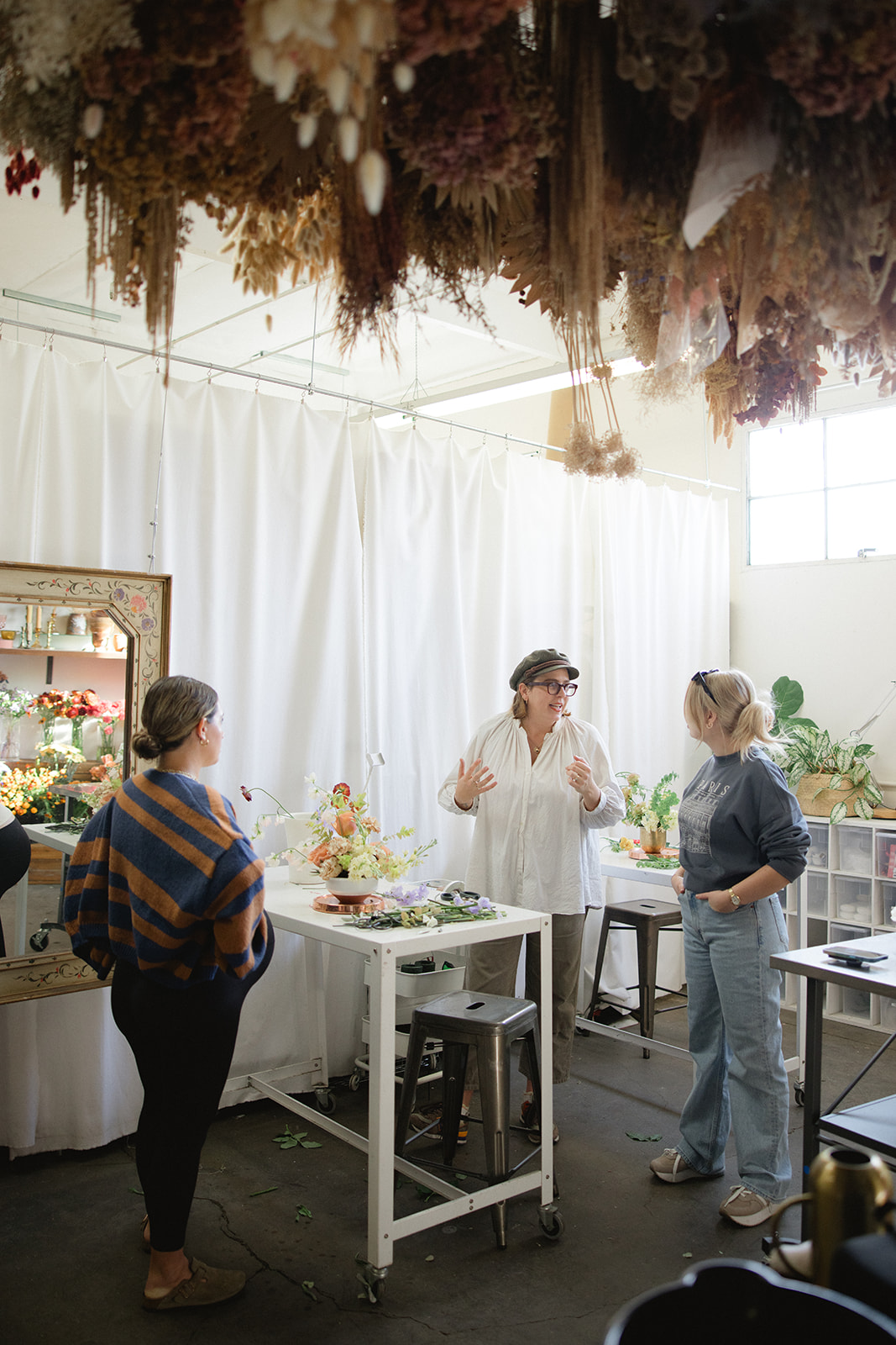 Three women in a bright room with white curtains, engaged in conversation near tables with floral arrangements.