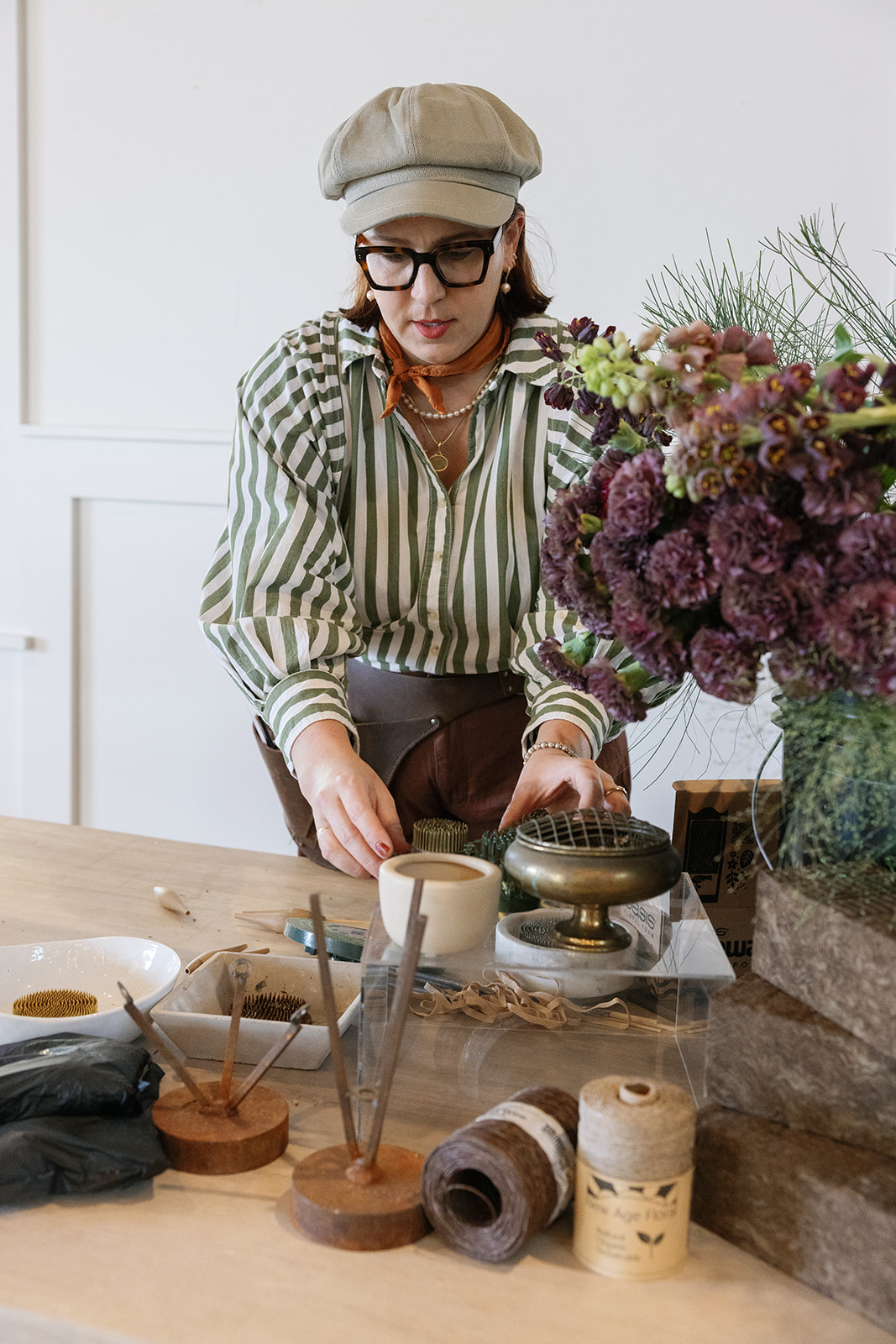 Woman wearing glasses, a beige hat, and striped shirt arranging items on a table with flowers and craft supplies.