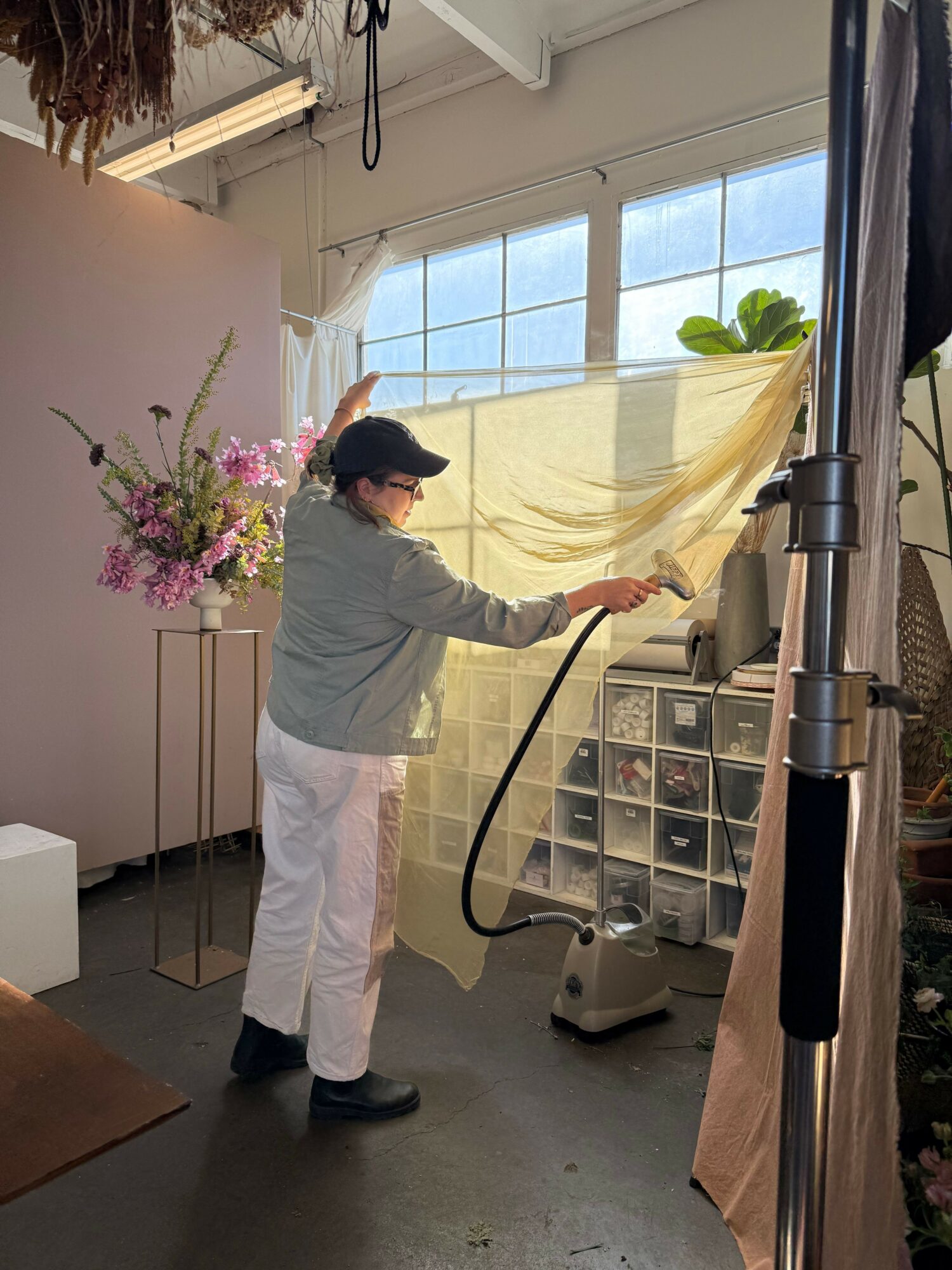 Child reaching up to adjust a yellow fabric near a window with sunlight, surrounded by flowers and storage drawers.