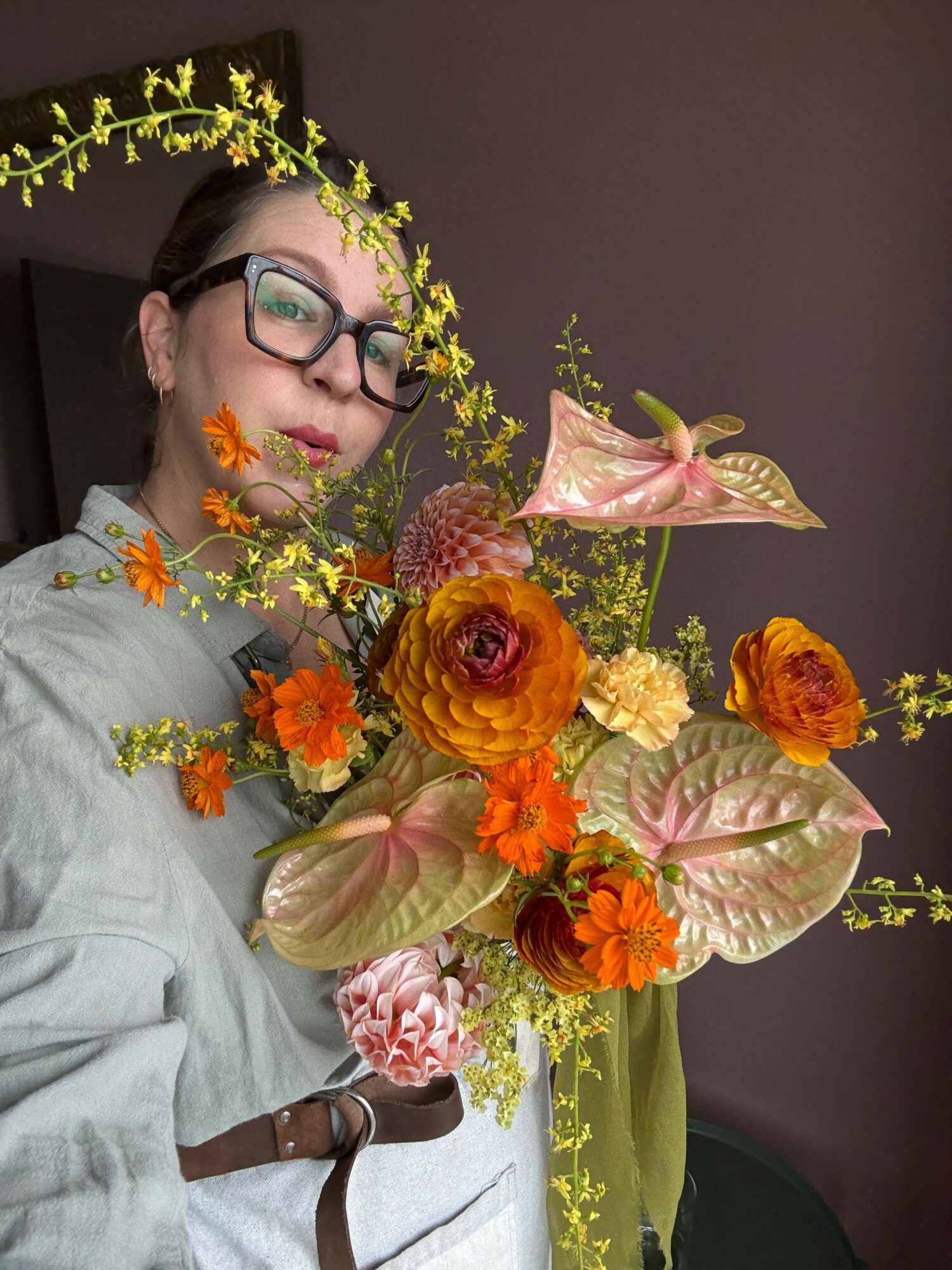 Person with glasses lying down, surrounded by a large bouquet of orange, pink, and cream flowers with green foliage.