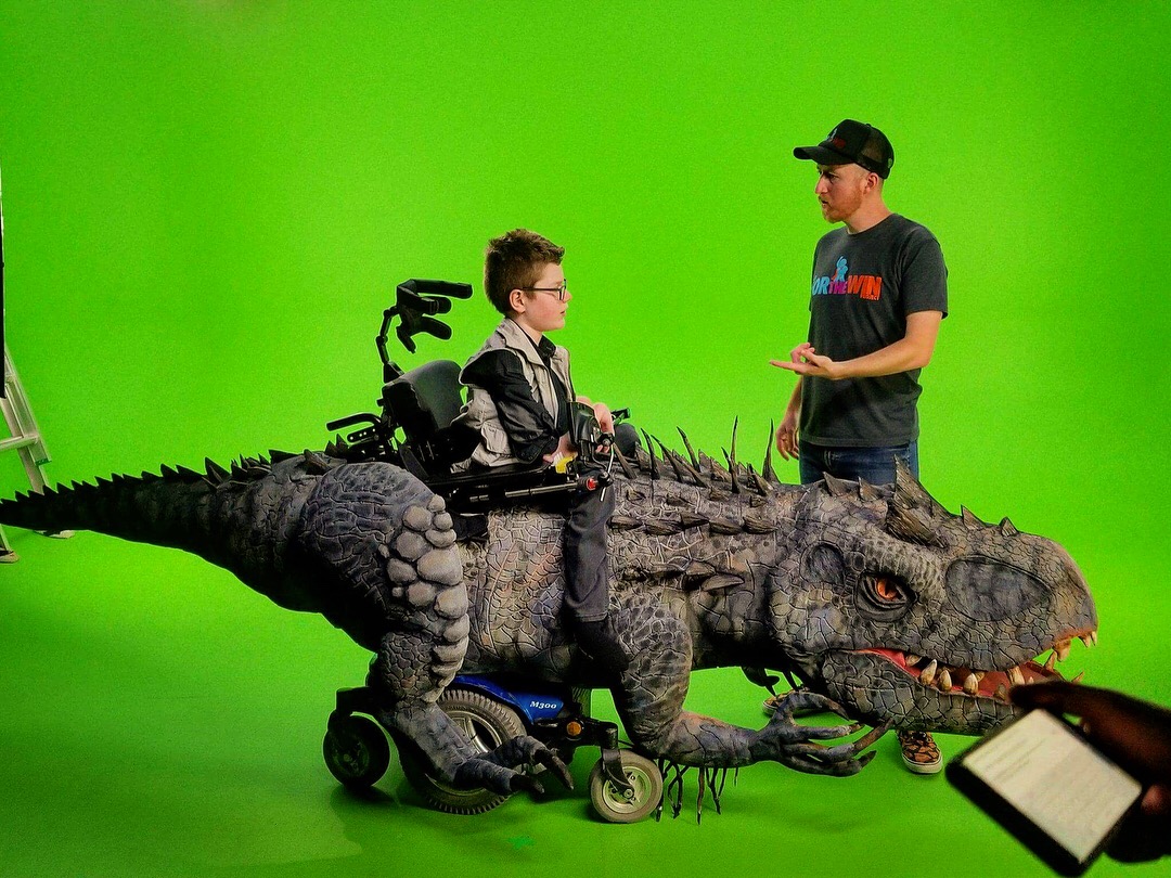 Child in a wheelchair with a dinosaur costume, talking to an adult in front of a green screen, tablet visible.