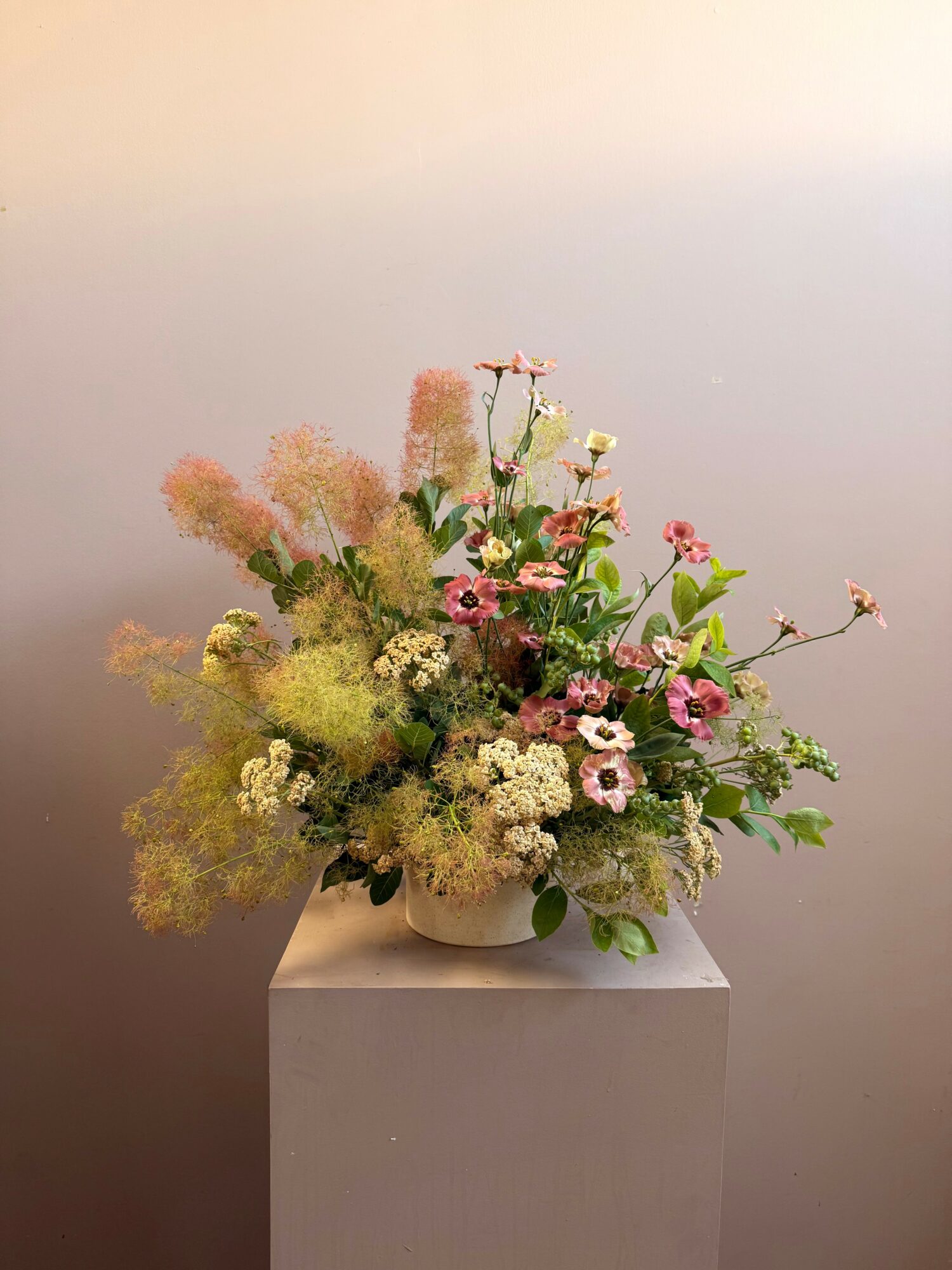 Flower arrangement with pink, white, and green flowers in a white vase on a pedestal against a plain background.