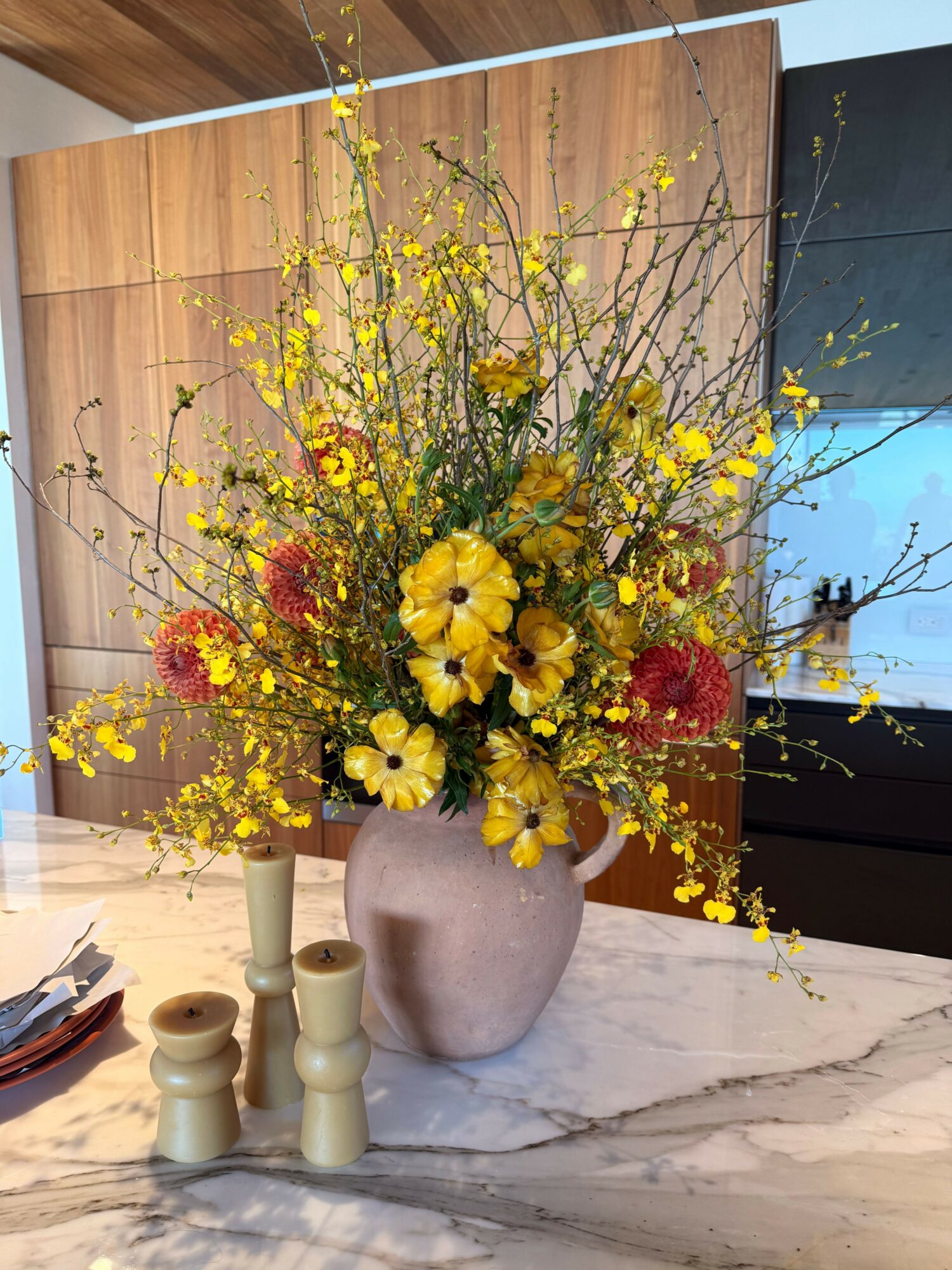 Vase with yellow, red, and green flowers on a marble surface, with candles nearby.