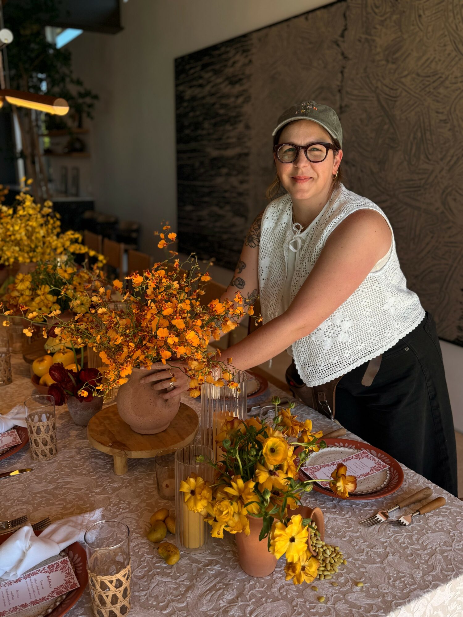 Woman with glasses and a hat arranging yellow flowers on a table, indoors with a dark wall background.