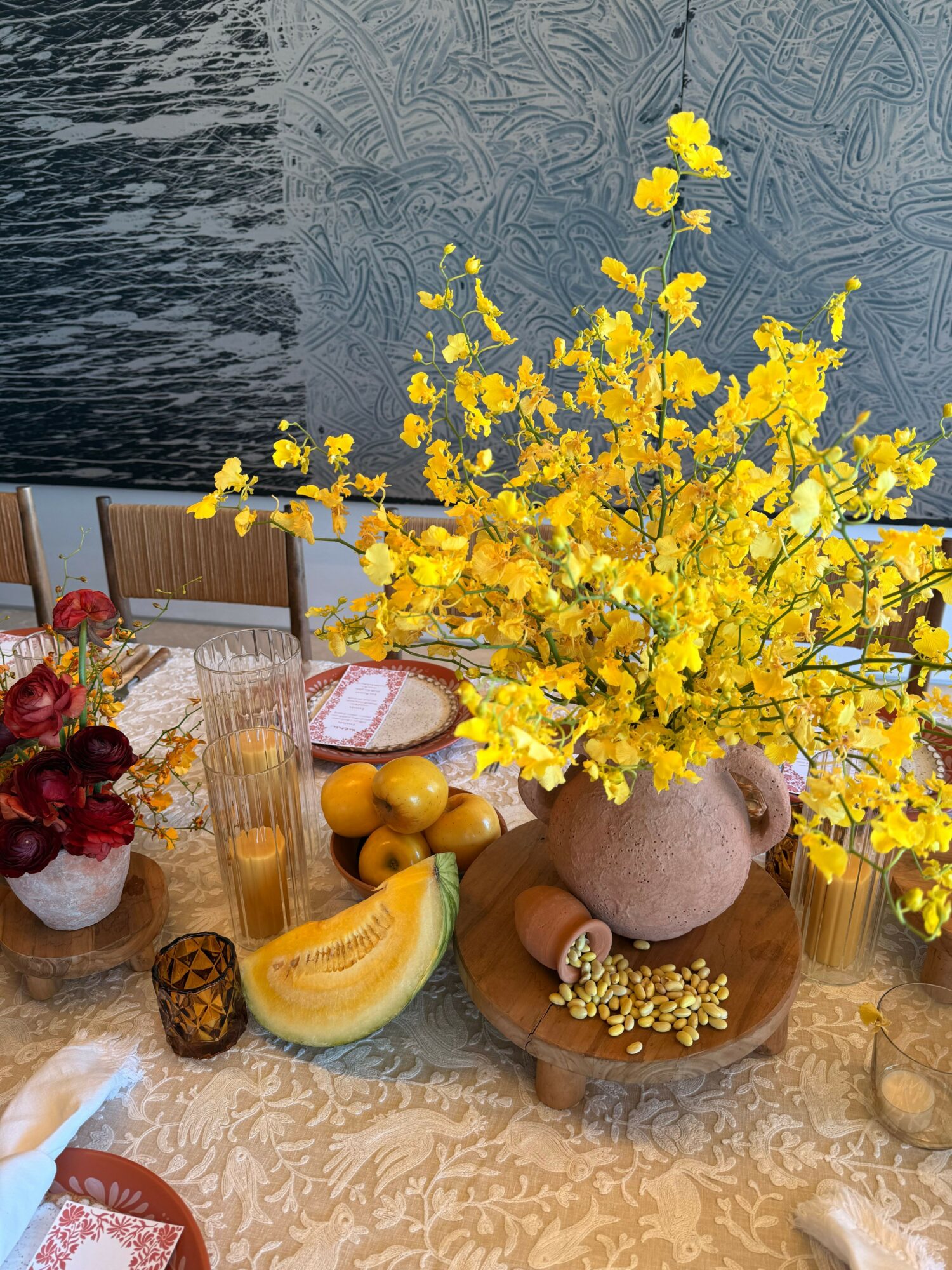 Yellow flowers in a vase, with fruits and decorative items on a table, viewed from above.