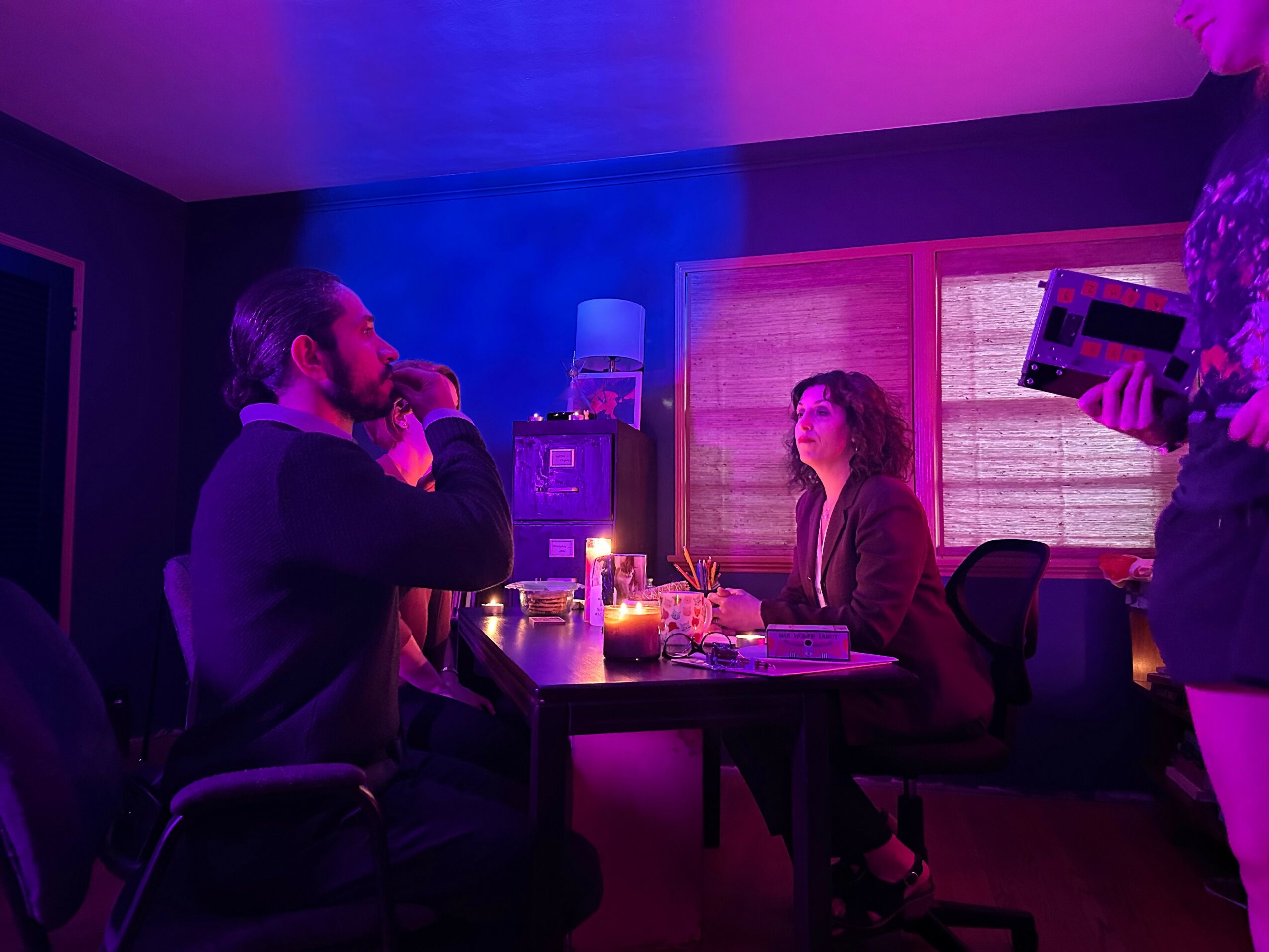 Two people sit at a table in a dimly lit room with purple and pink lighting, candles, and a window with blinds.