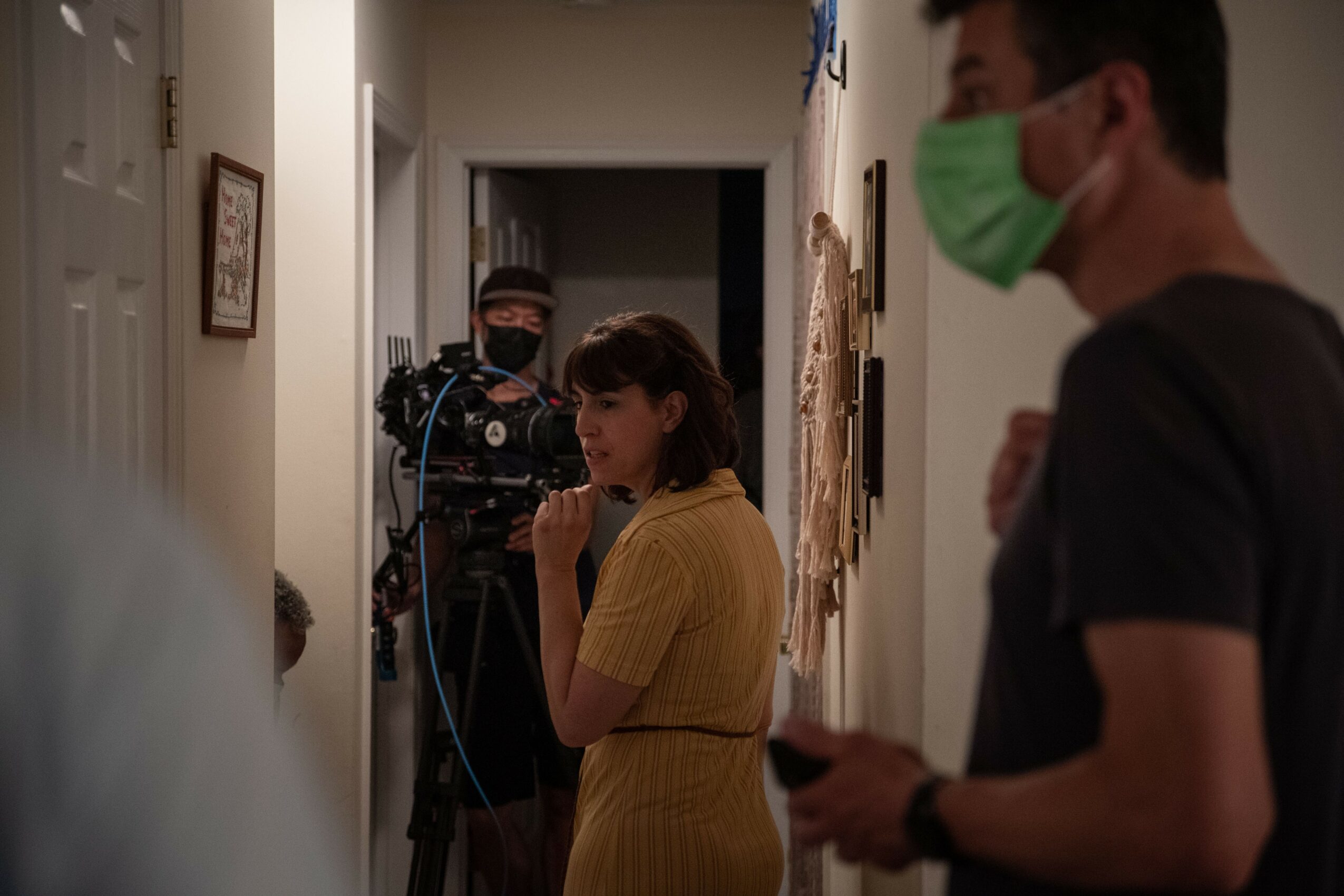 People in masks filming in a hallway, woman in yellow dress, man with phone, camera operator in background, indoor setting