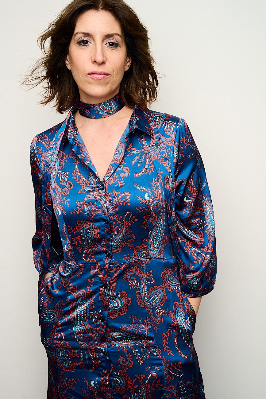 Woman with shoulder-length wavy hair wearing a blue patterned silk shirt and choker necklace, standing against a plain background.