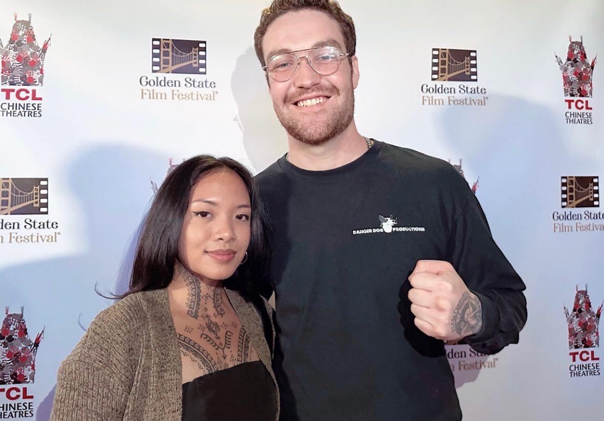 Two people standing together, smiling, in front of a backdrop with logos, one with glasses and a beard, the other with long dark hair.