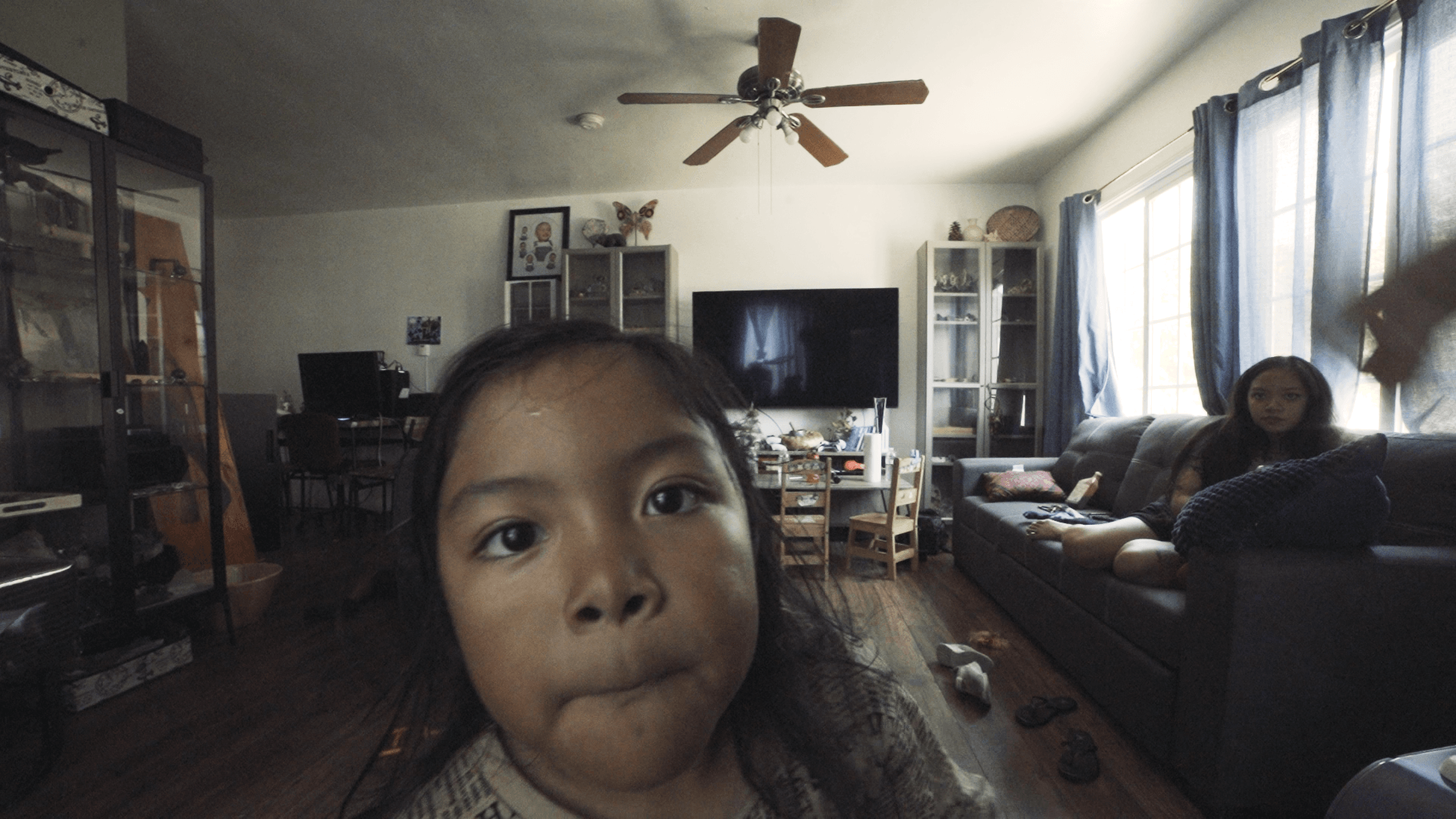Child with dark hair looking at camera in living room with TV, sofa, and windows with curtains