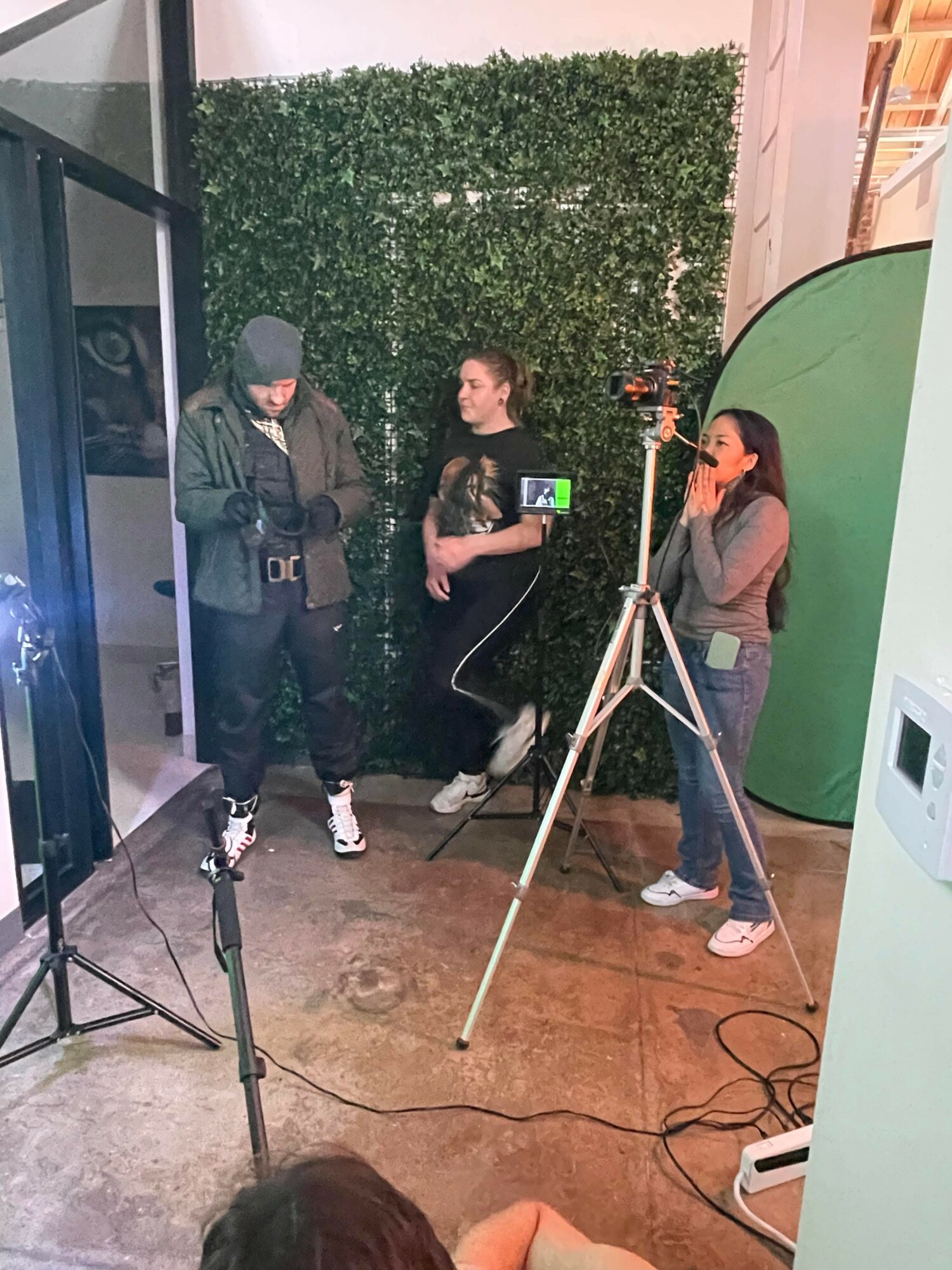 Three people are in a photo studio with a green backdrop, one person is being interviewed, and two others are recording.
