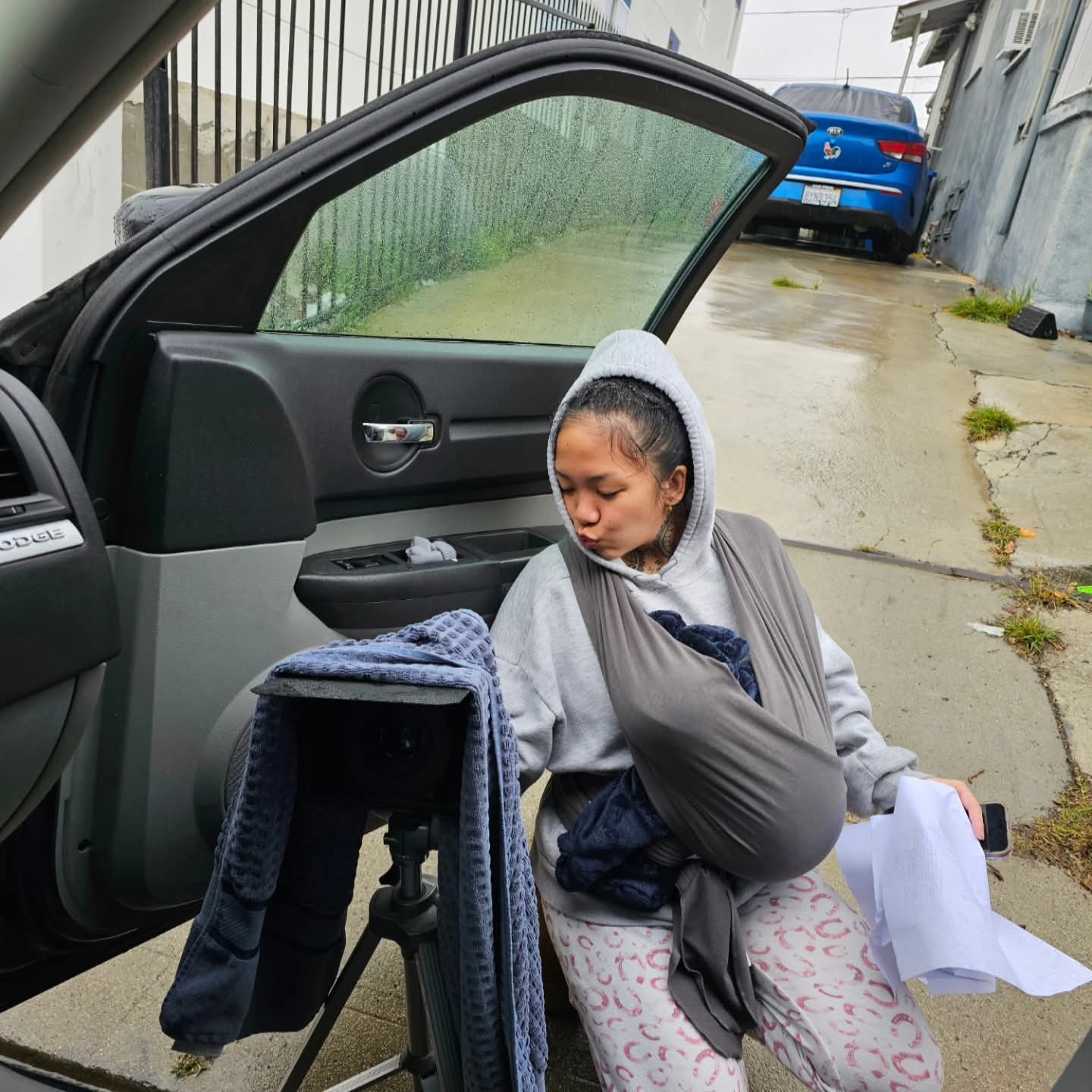 Young woman sitting on the ground outside a car, holding a paper, with a camera on a tripod nearby.