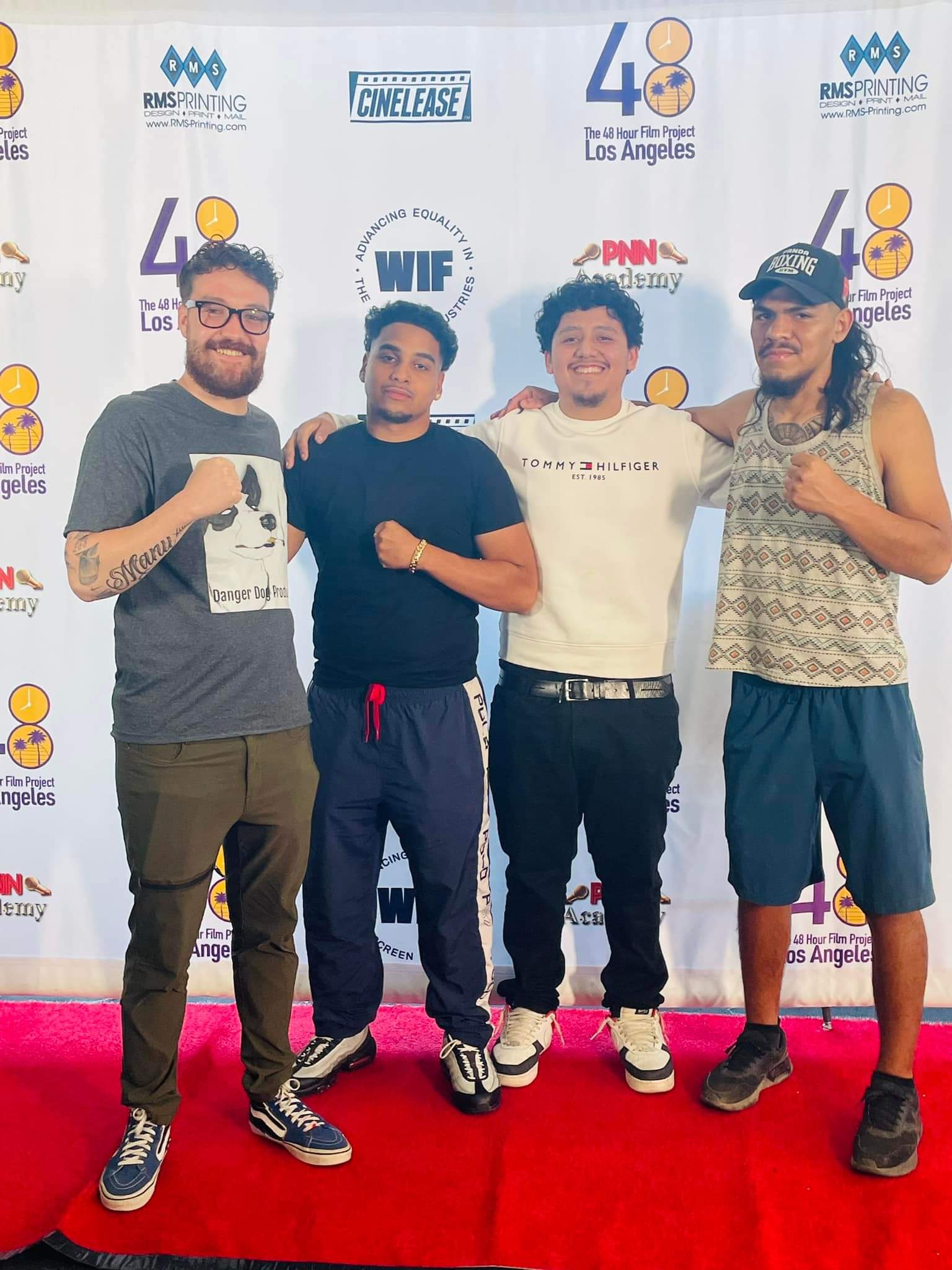 Four young men standing on a red carpet in front of a backdrop with logos, smiling and posing for the photo.