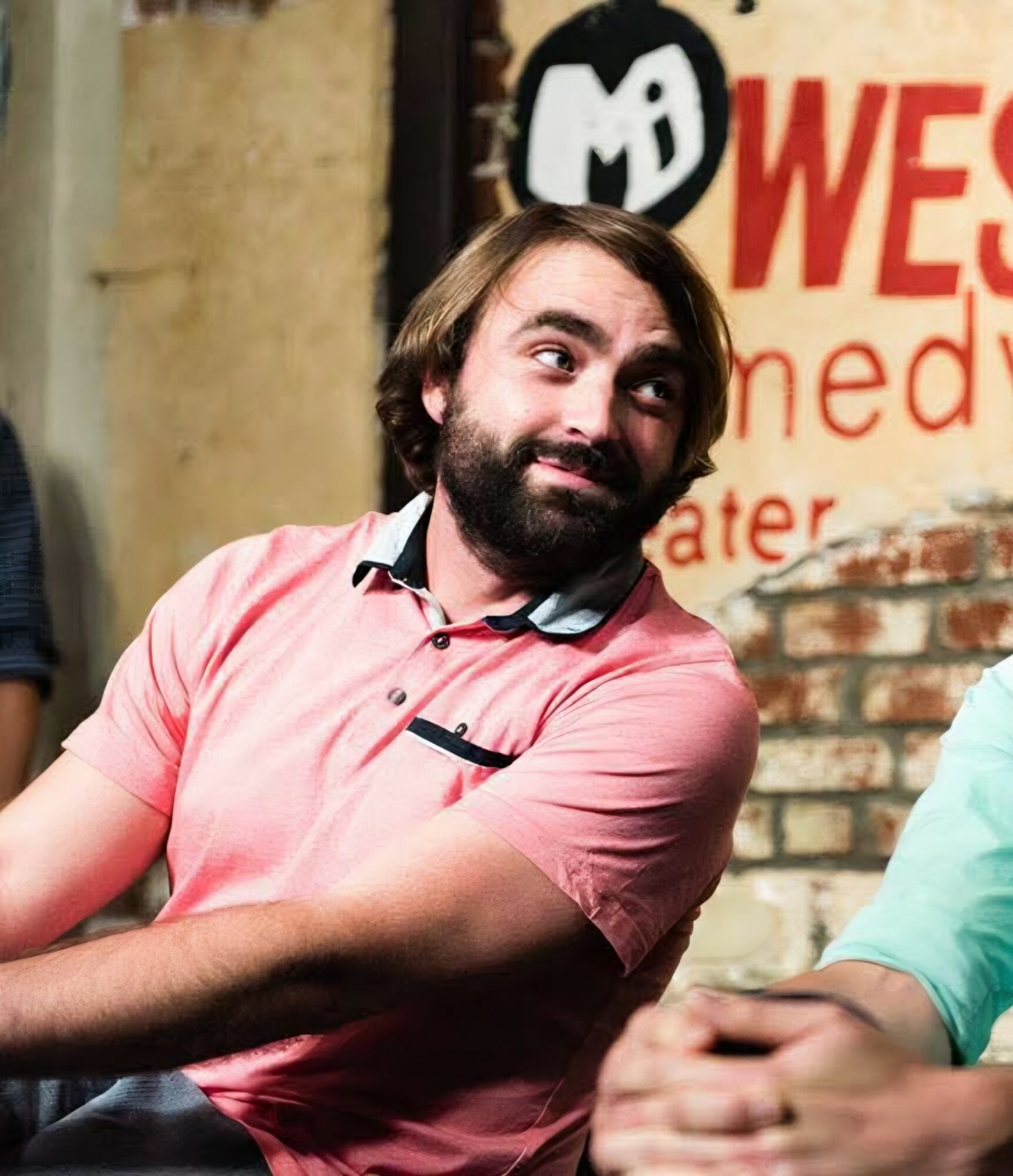 Man with beard and long hair smiling, wearing a pink shirt, in a comedy club setting.