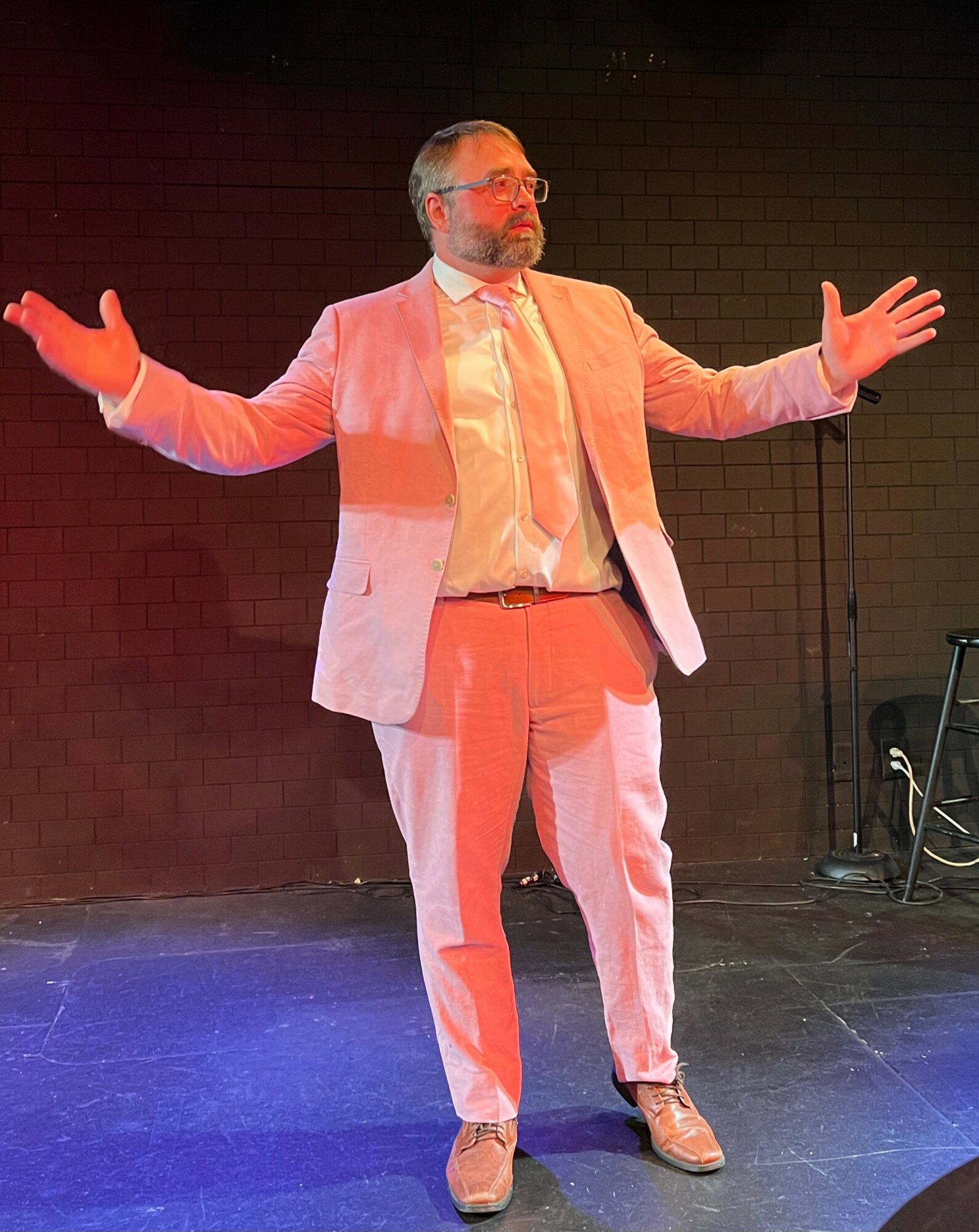 Man in light-colored suit with arms outstretched standing on stage against dark background.