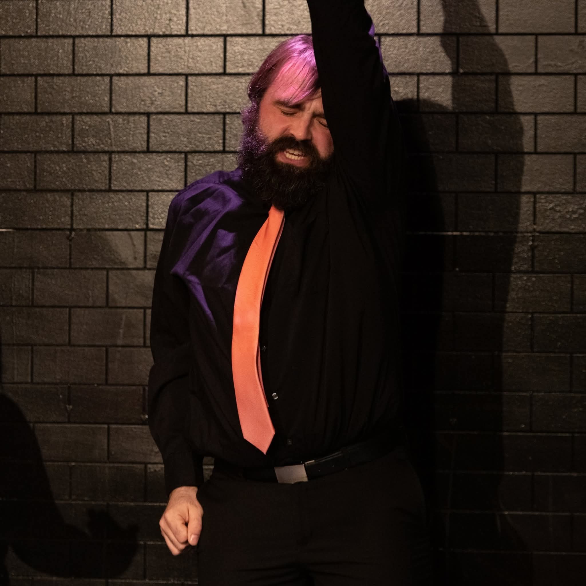 Person with beard and pink hair, wearing a black shirt and orange tie, standing against a brick wall with arm raised.