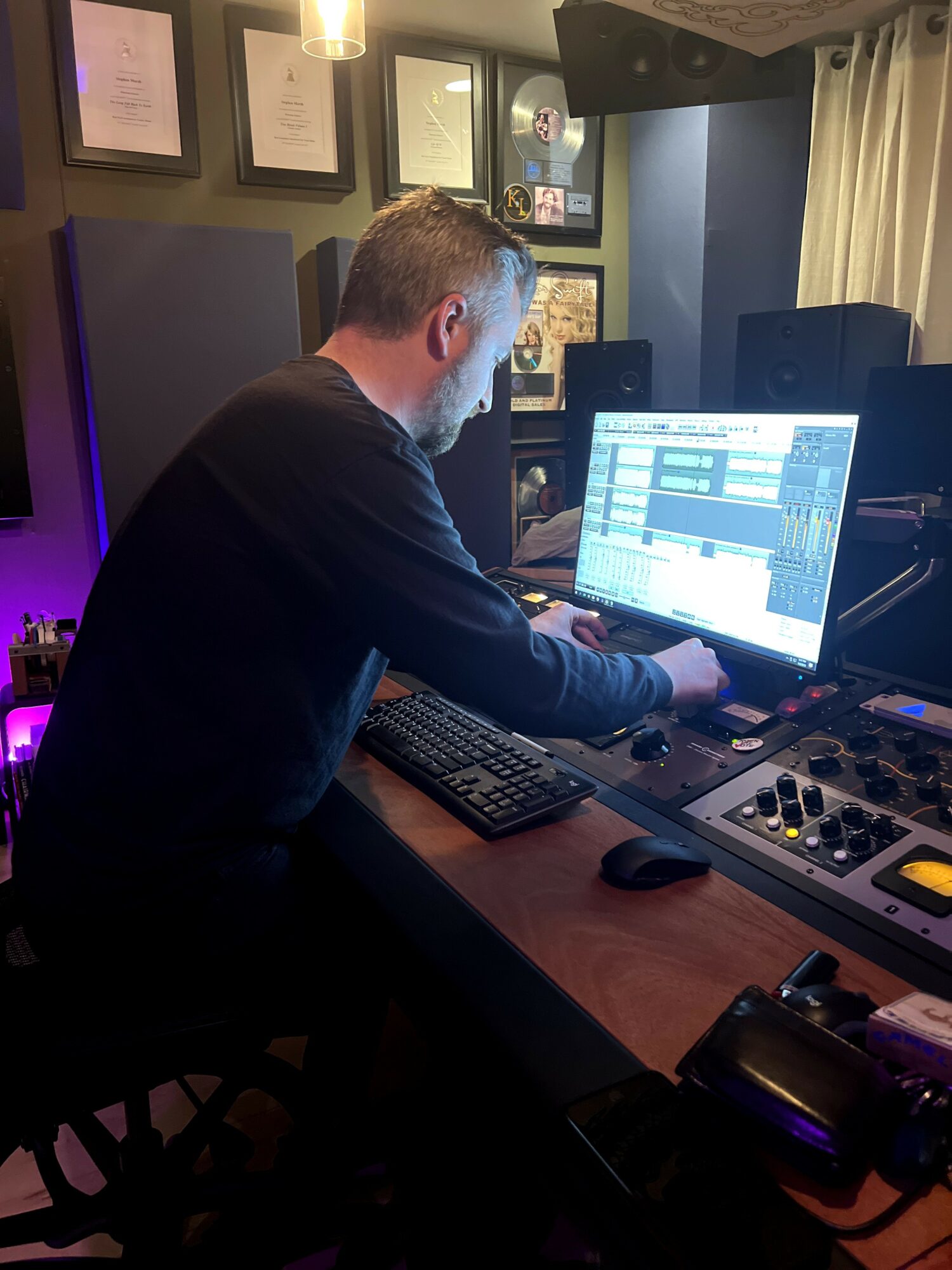 Person working at a music production setup with a computer, keyboard, and audio equipment in a room with framed certificates on the wall.