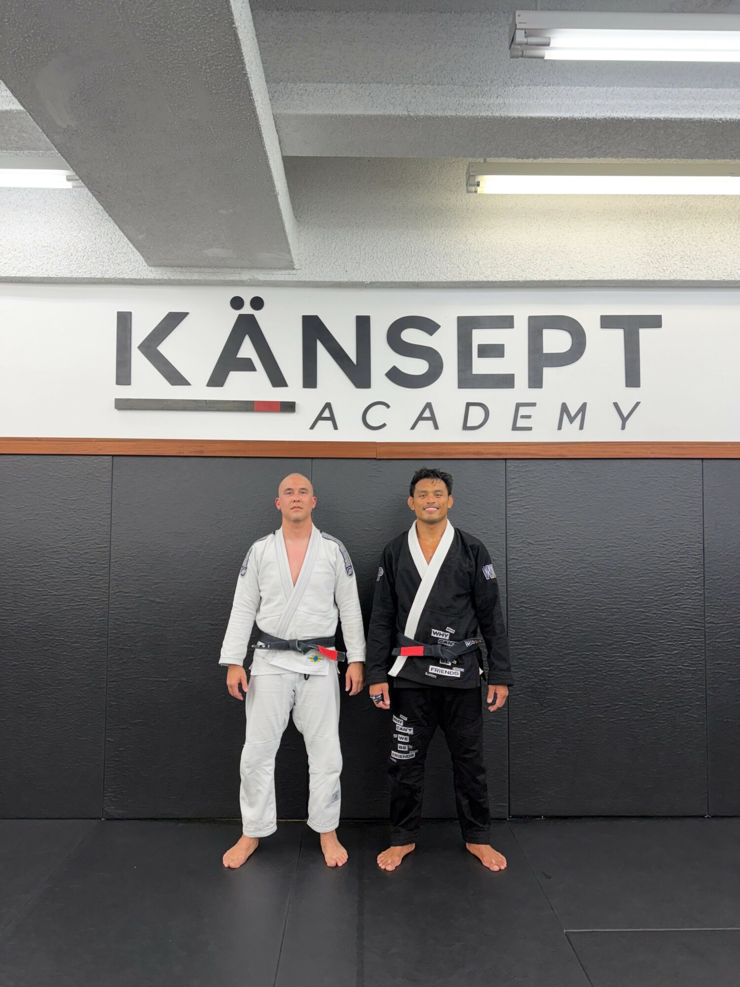 Two martial artists in gi uniforms standing in front of a sign that reads 'KANSÉPT ACADEMY'.
