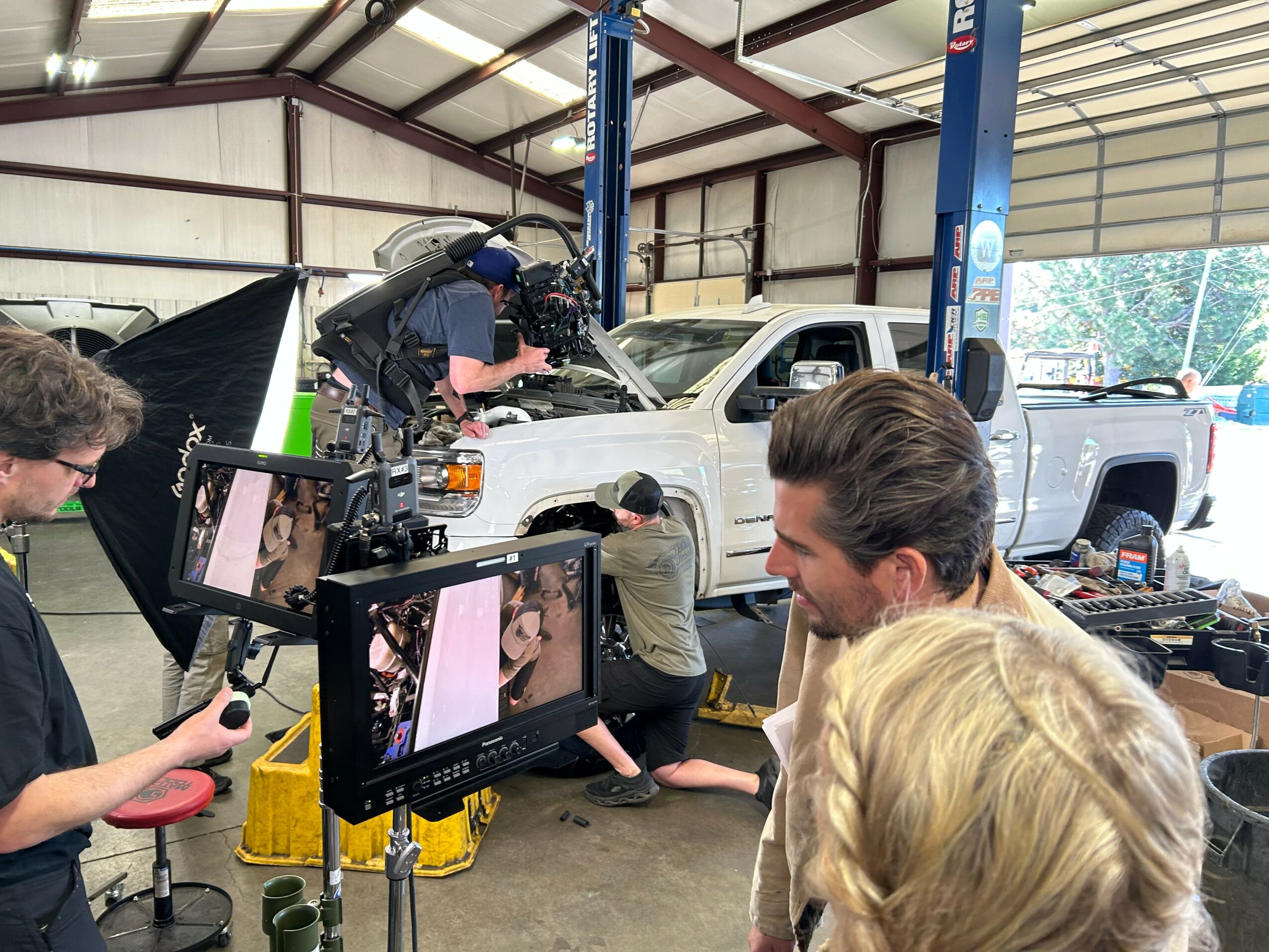 People working on a white pickup truck inside a garage, with cameras and equipment set up around them.