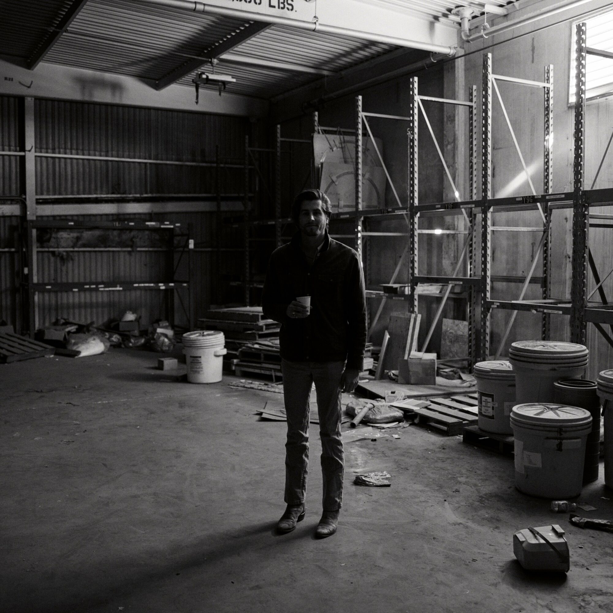 Person standing in a warehouse with shelves and buckets, holding a phone, in black and white.
