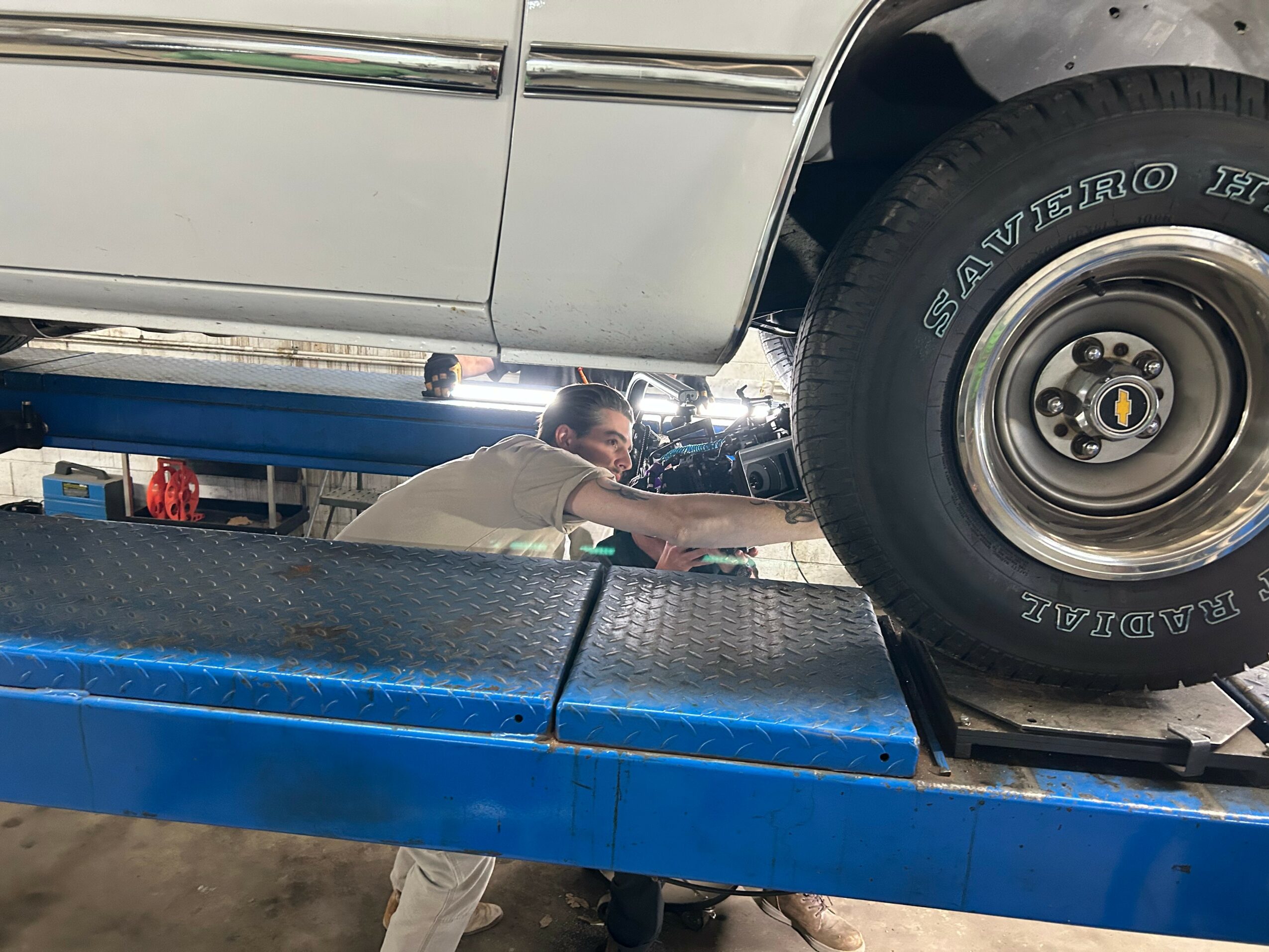 Person working underneath a vehicle on a blue lift, with the vehicle's wheel visible.