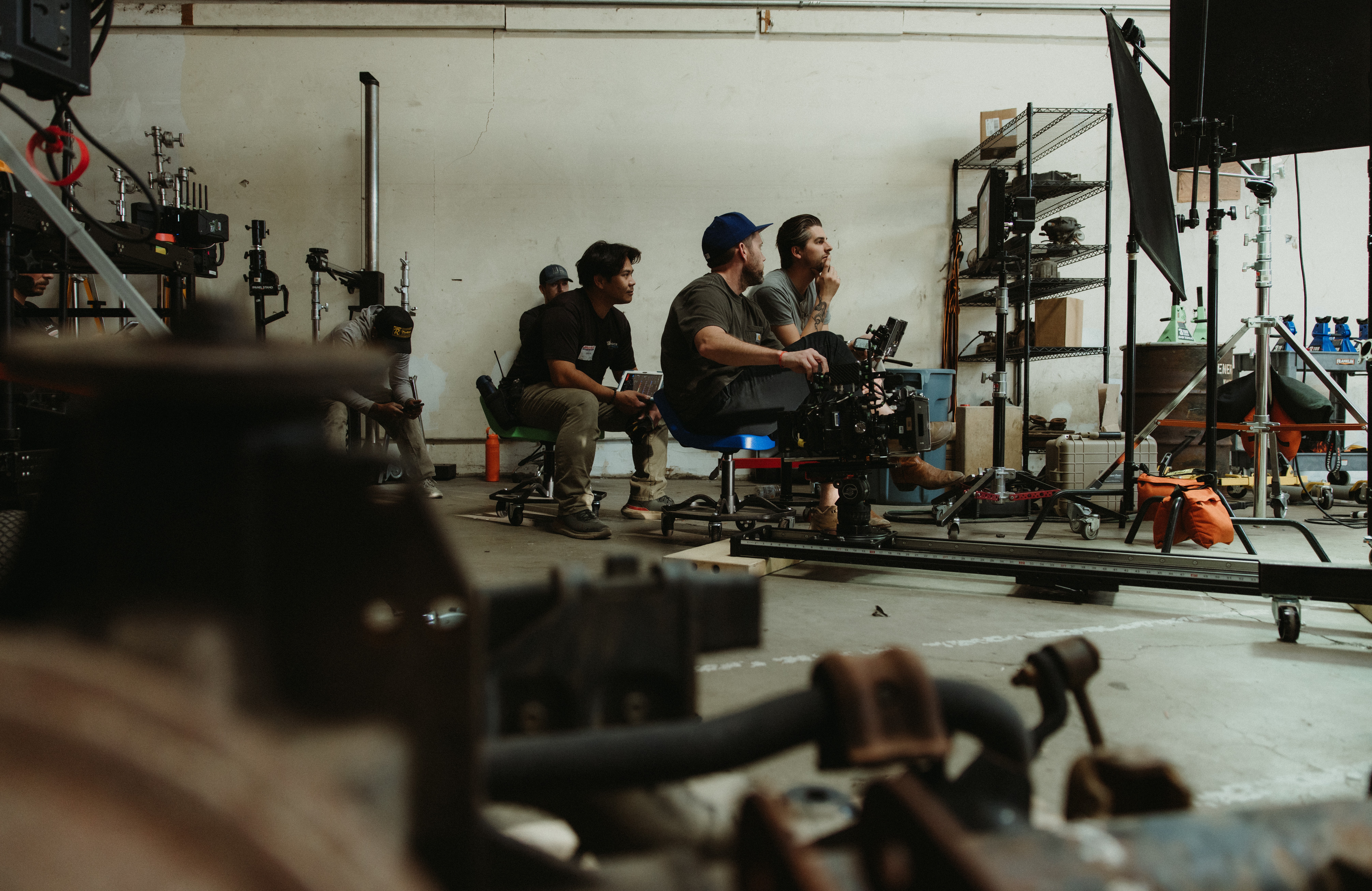 Group of people working with camera equipment in a studio or warehouse space, with shelves and equipment in the background.