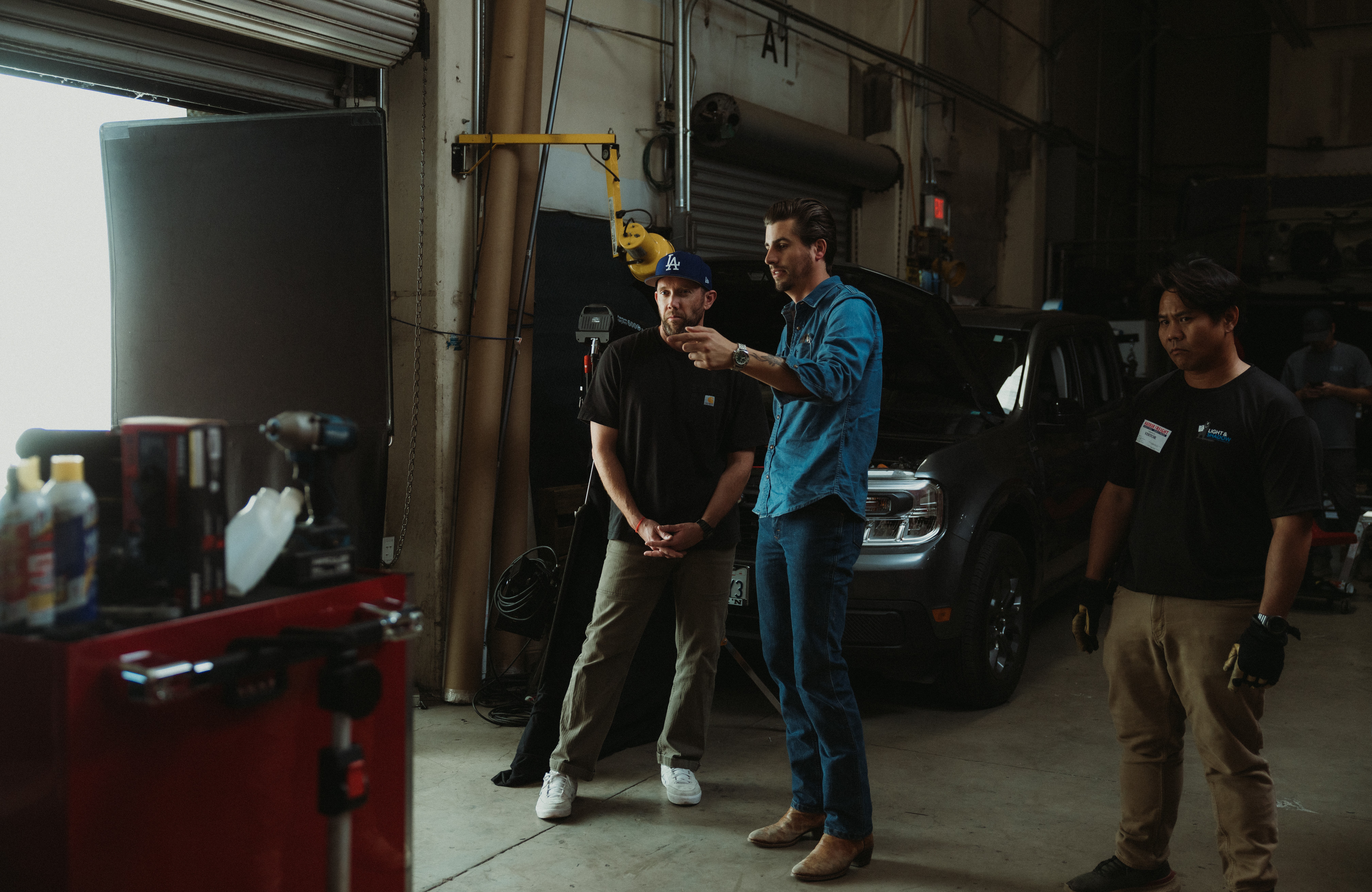 Three people standing and talking inside a garage or warehouse, with a vehicle and equipment visible in the background.