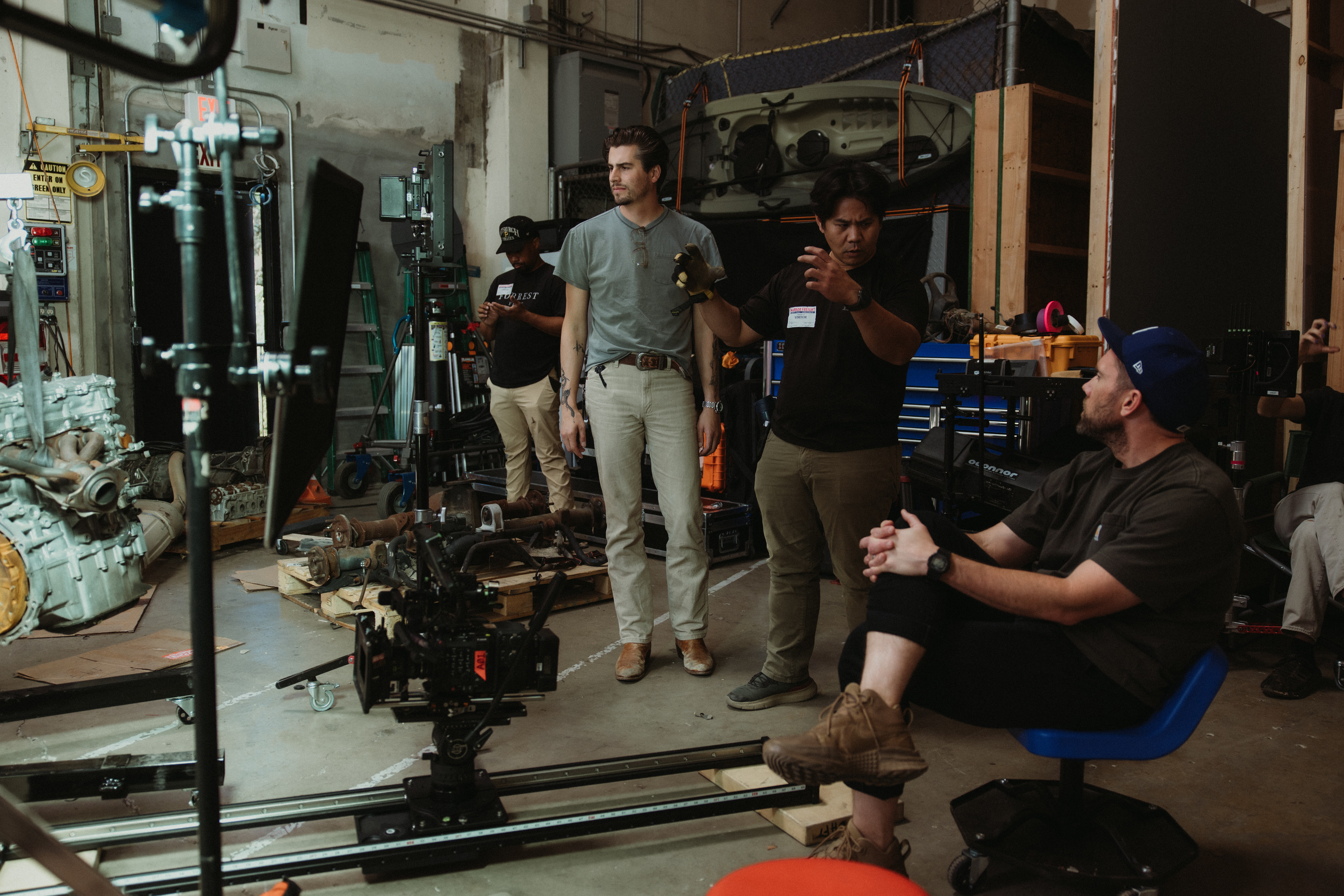 Group of people in a workshop with equipment and machinery, some sitting and others standing, engaged in conversation.