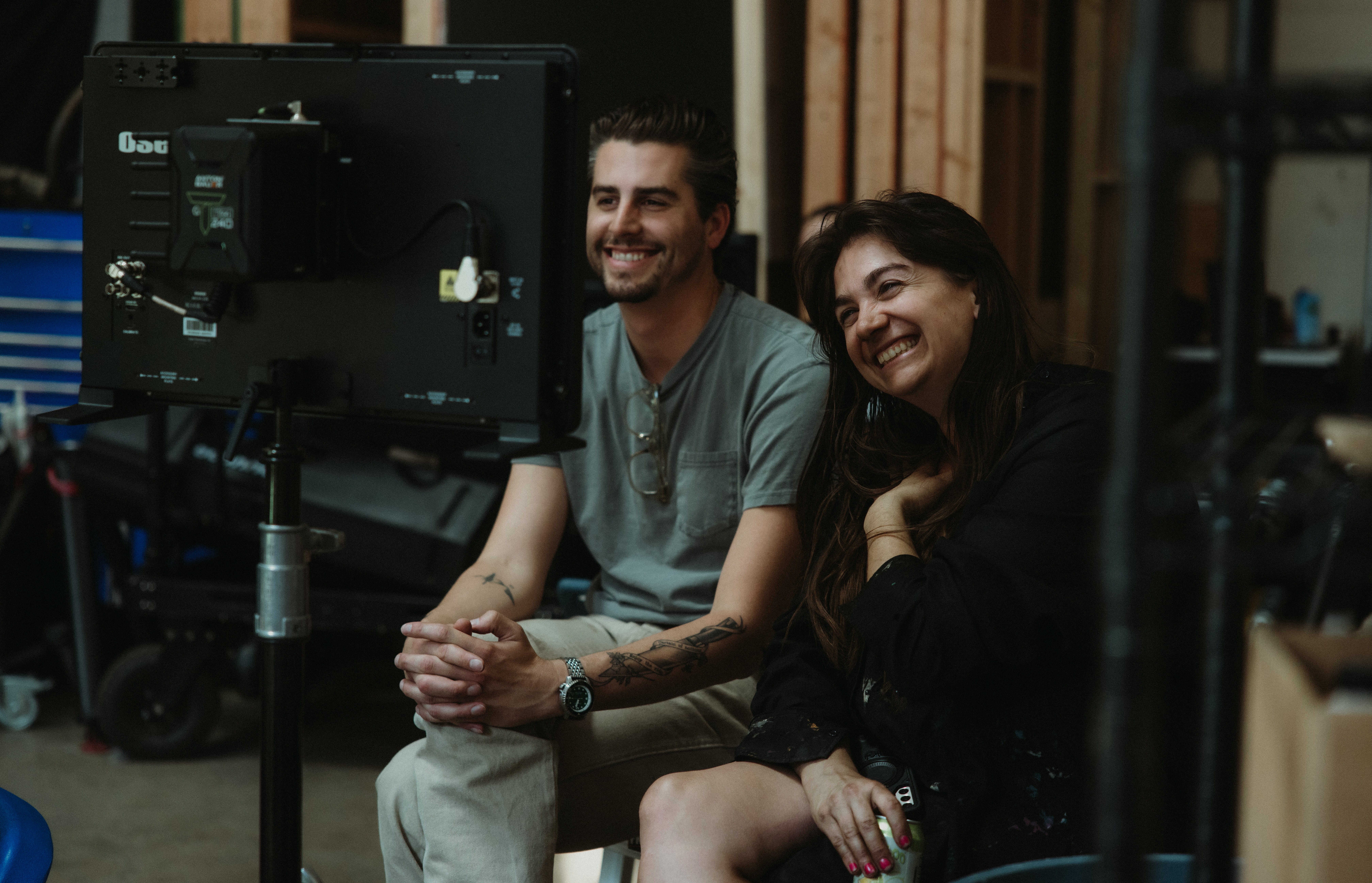 Two people sitting and smiling in a room with a computer monitor, surrounded by equipment and wooden walls.