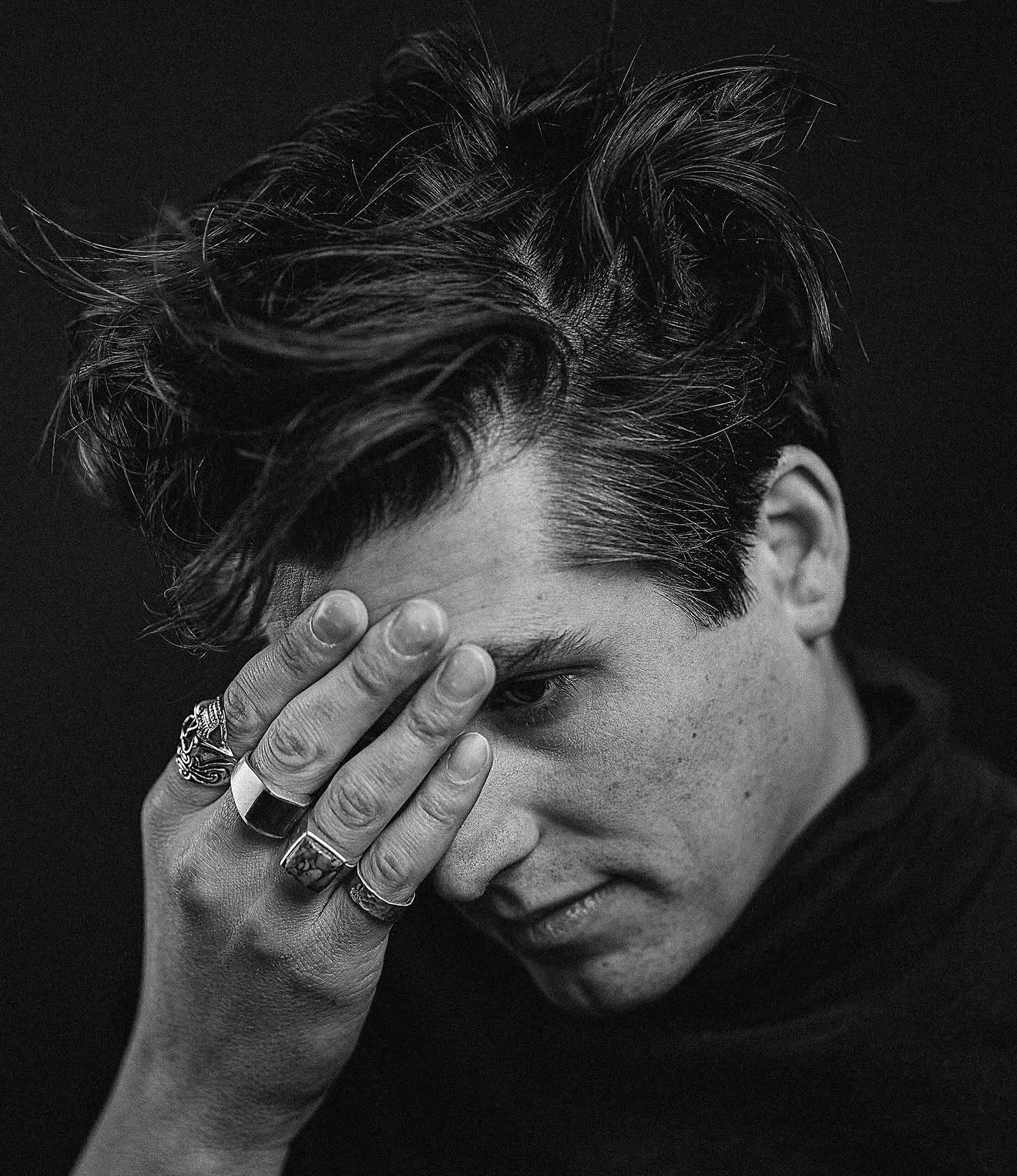 Young person with tousled hair covering part of face with hand, wearing rings, in black and white.