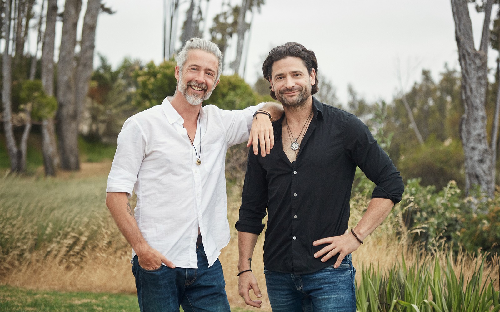 Two men standing outdoors, smiling, with trees and grass in the background.