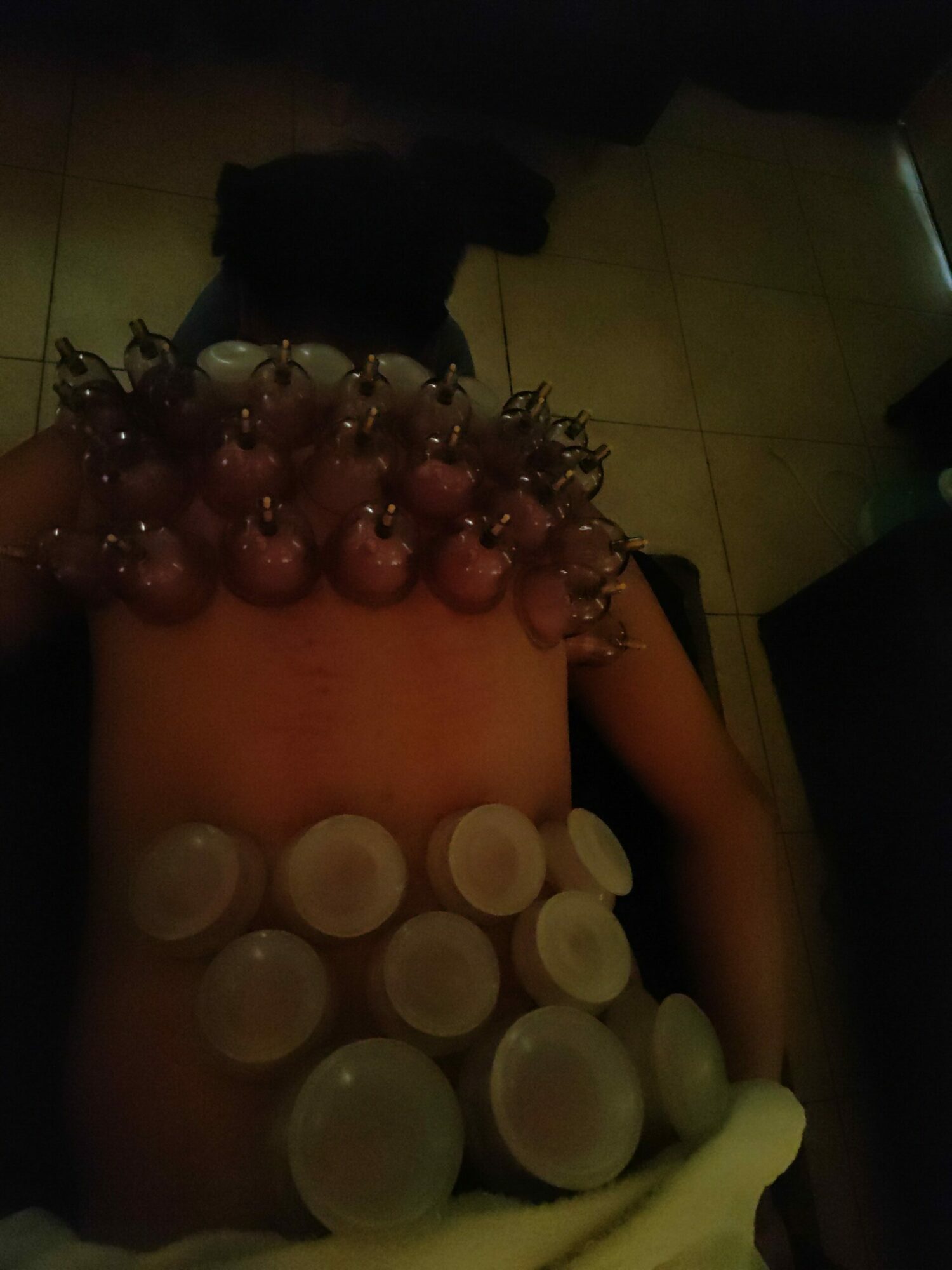 Person lying down with various objects on their stomach, including round containers and a beaded accessory, in a dimly lit room.