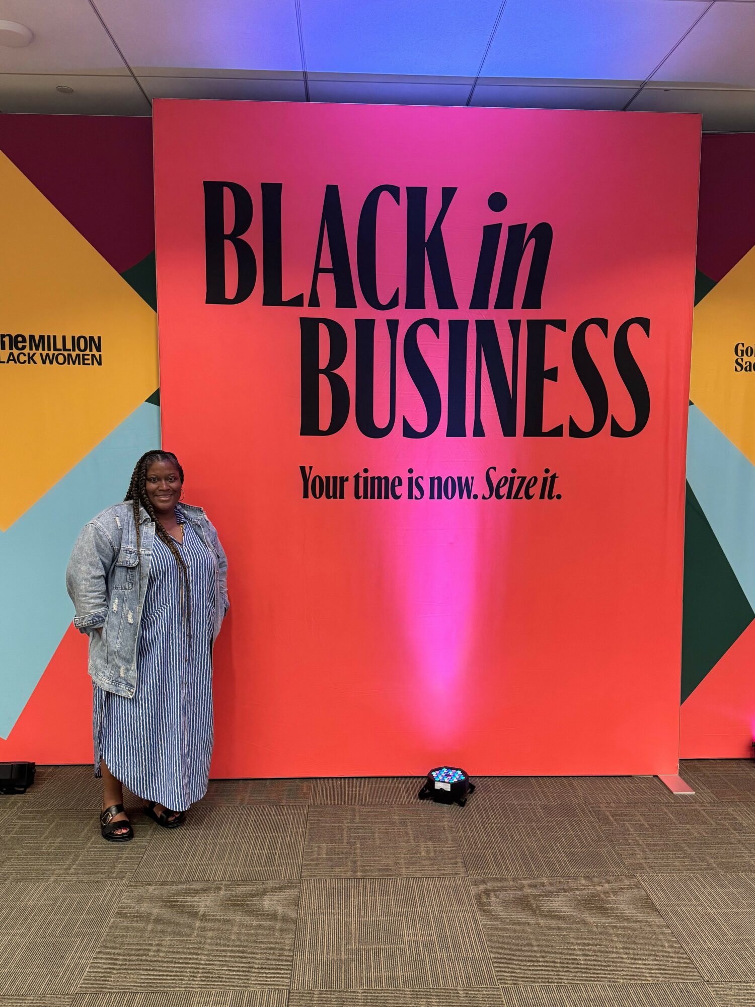 Person standing in front of a large red and pink display with text about Black in Business, wearing a gray coat and black shoes.