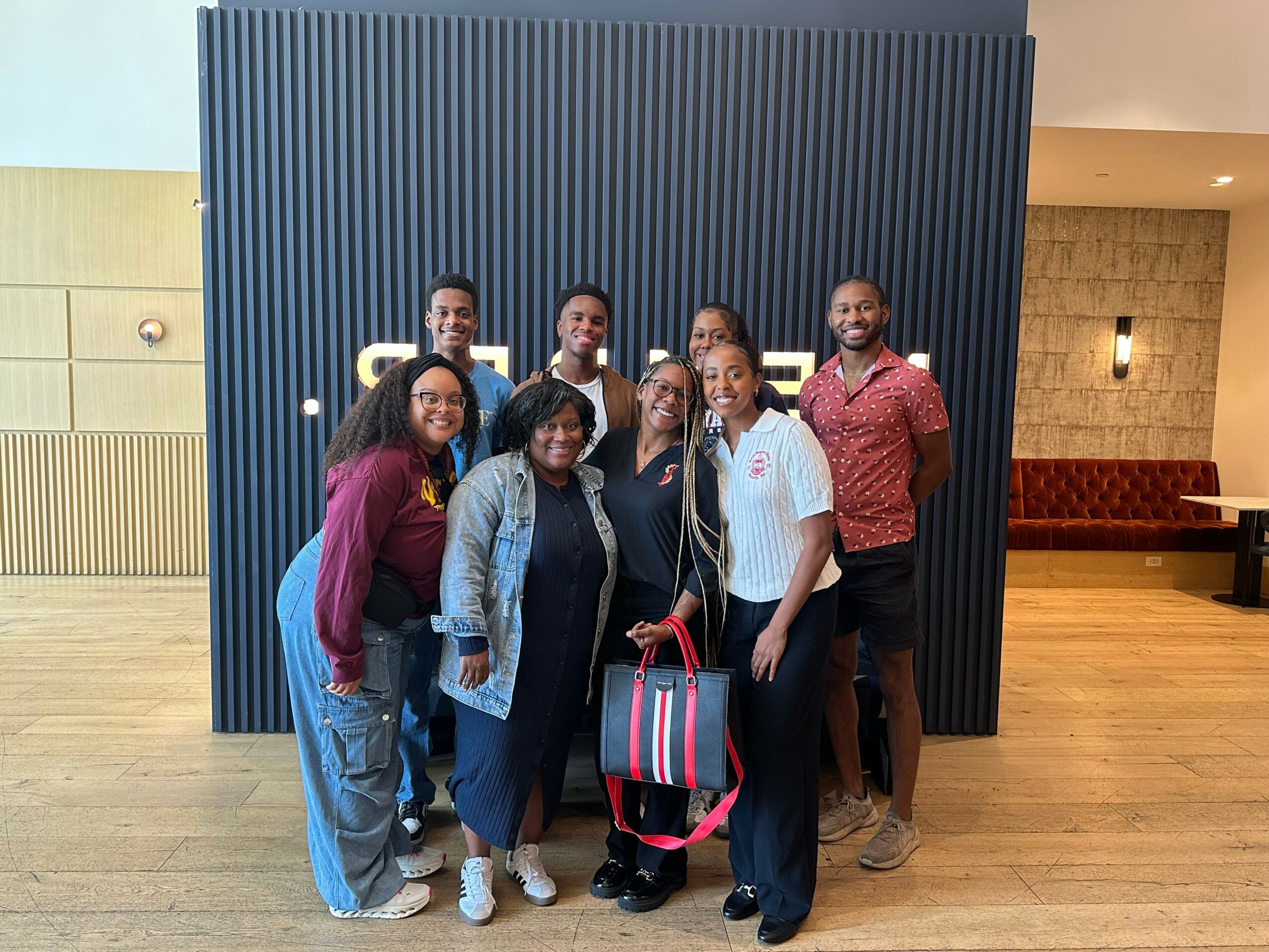 Group of nine people standing indoors in front of a black wall with vertical lines, smiling and posing for the photo.