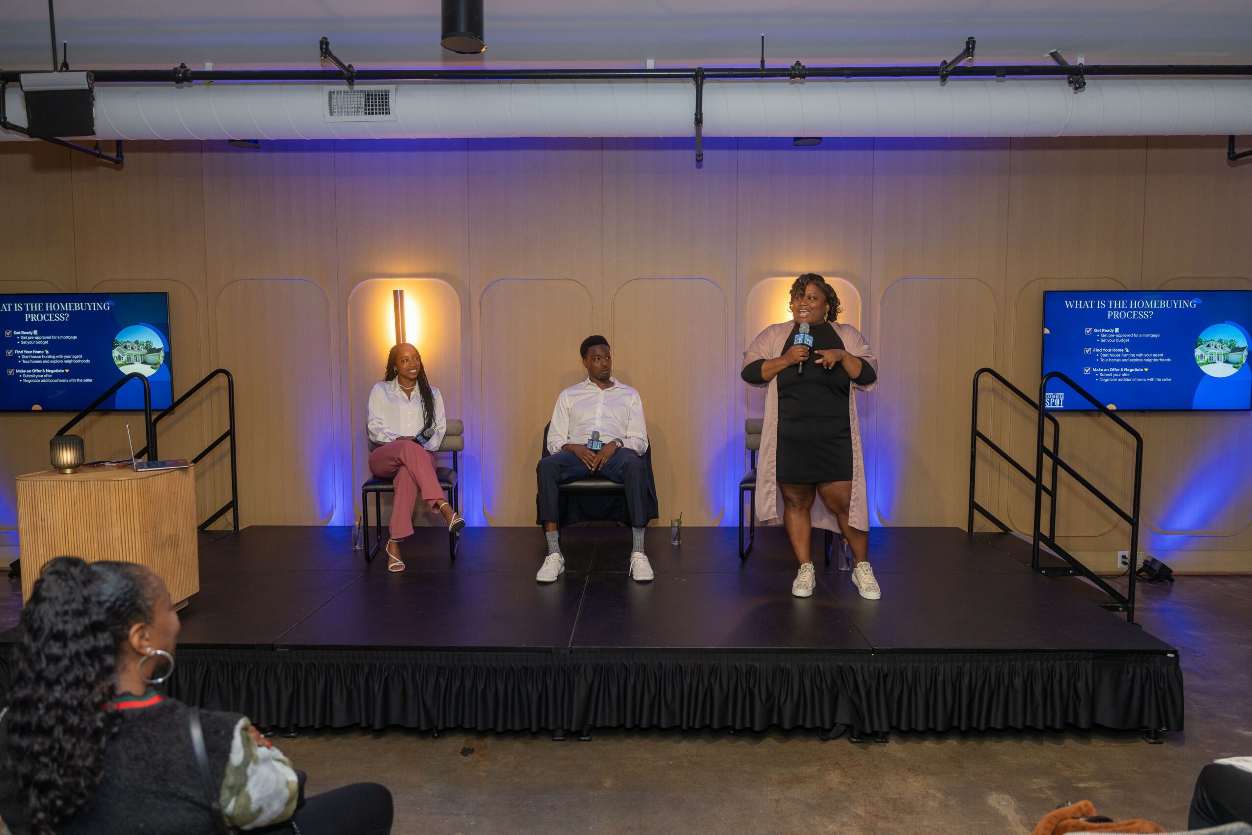 Three people on stage, one woman speaking into a microphone, two seated, two screens with presentation slides, audience visible.