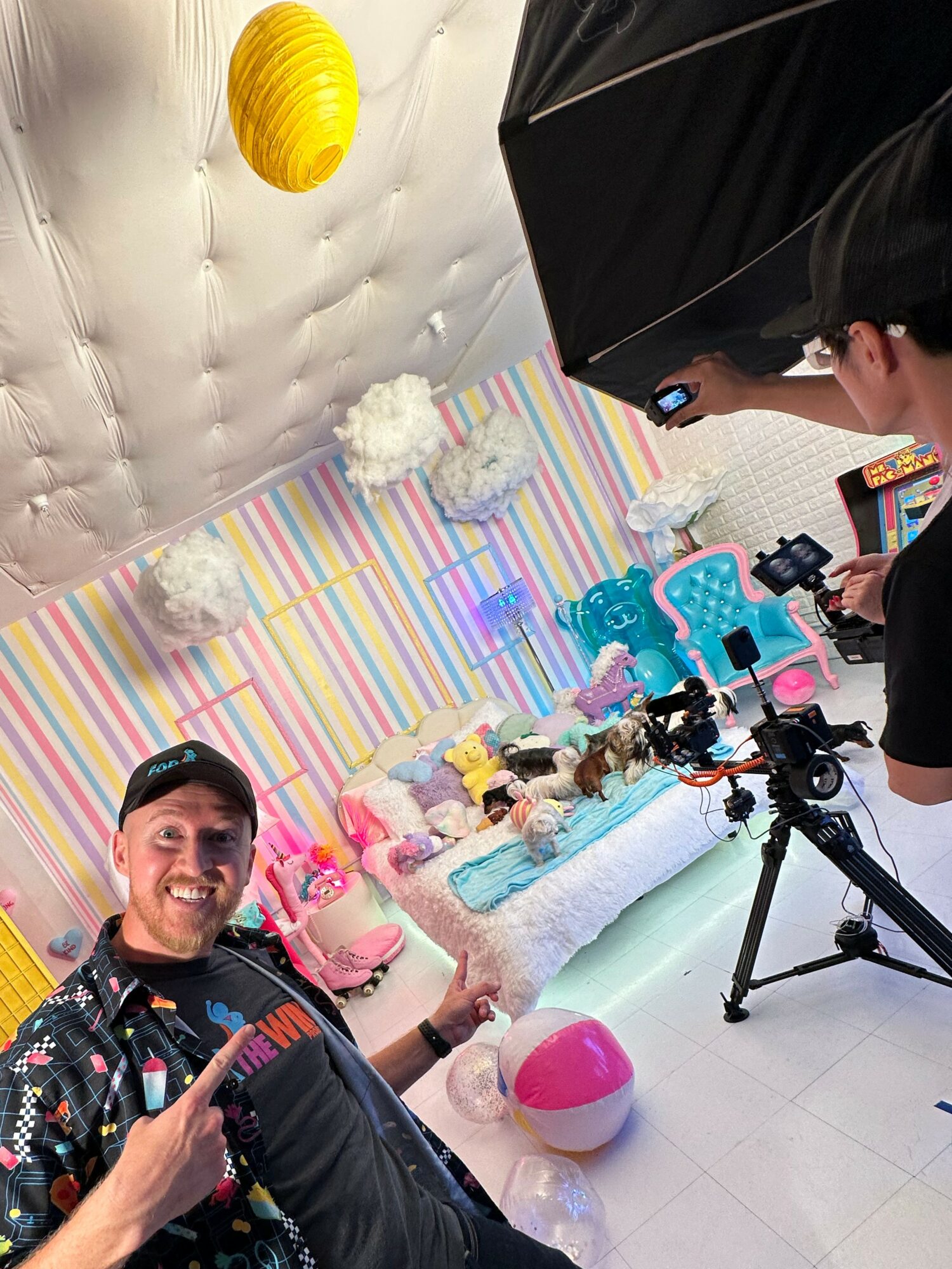 Man smiling and pointing at camera in colorful party room with striped wall, balloons, and camera equipment.