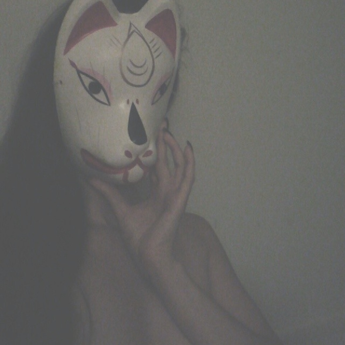 Person wearing a fox mask covering their face, with hand touching the mask, against a plain background.