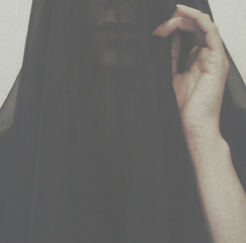 Person holding a sheer black fabric near face, hand visible, background plain, face partially obscured by fabric.