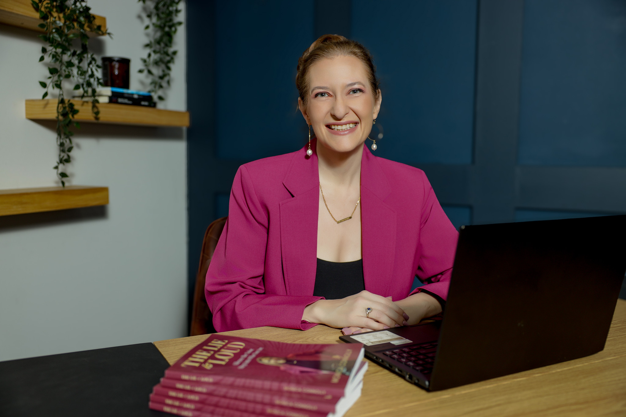 Woman in pink blazer sitting at desk with laptop, smiling, in a modern office setting.