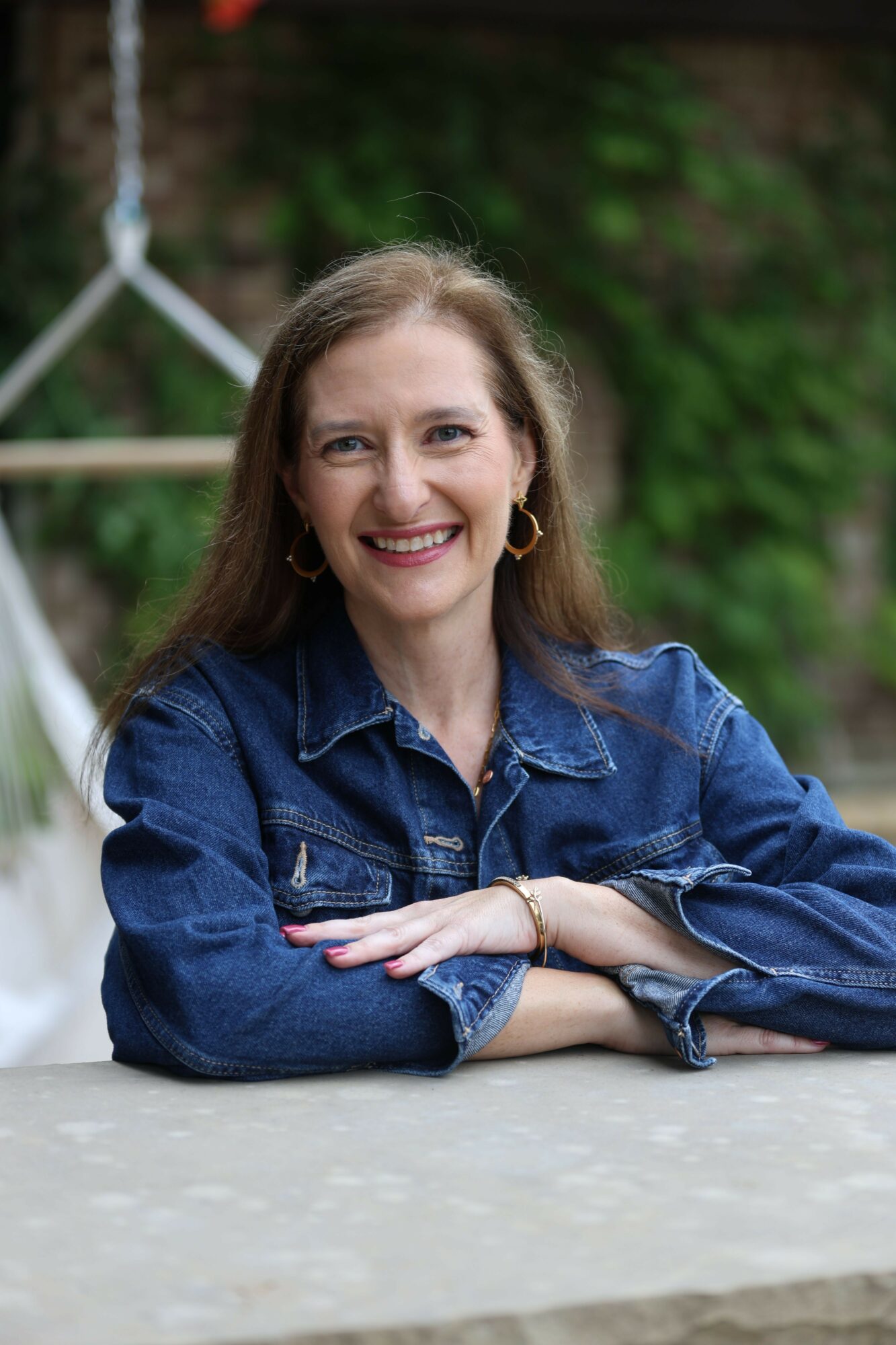 Woman with long hair smiling, wearing a denim jacket, sitting outdoors with greenery in the background.