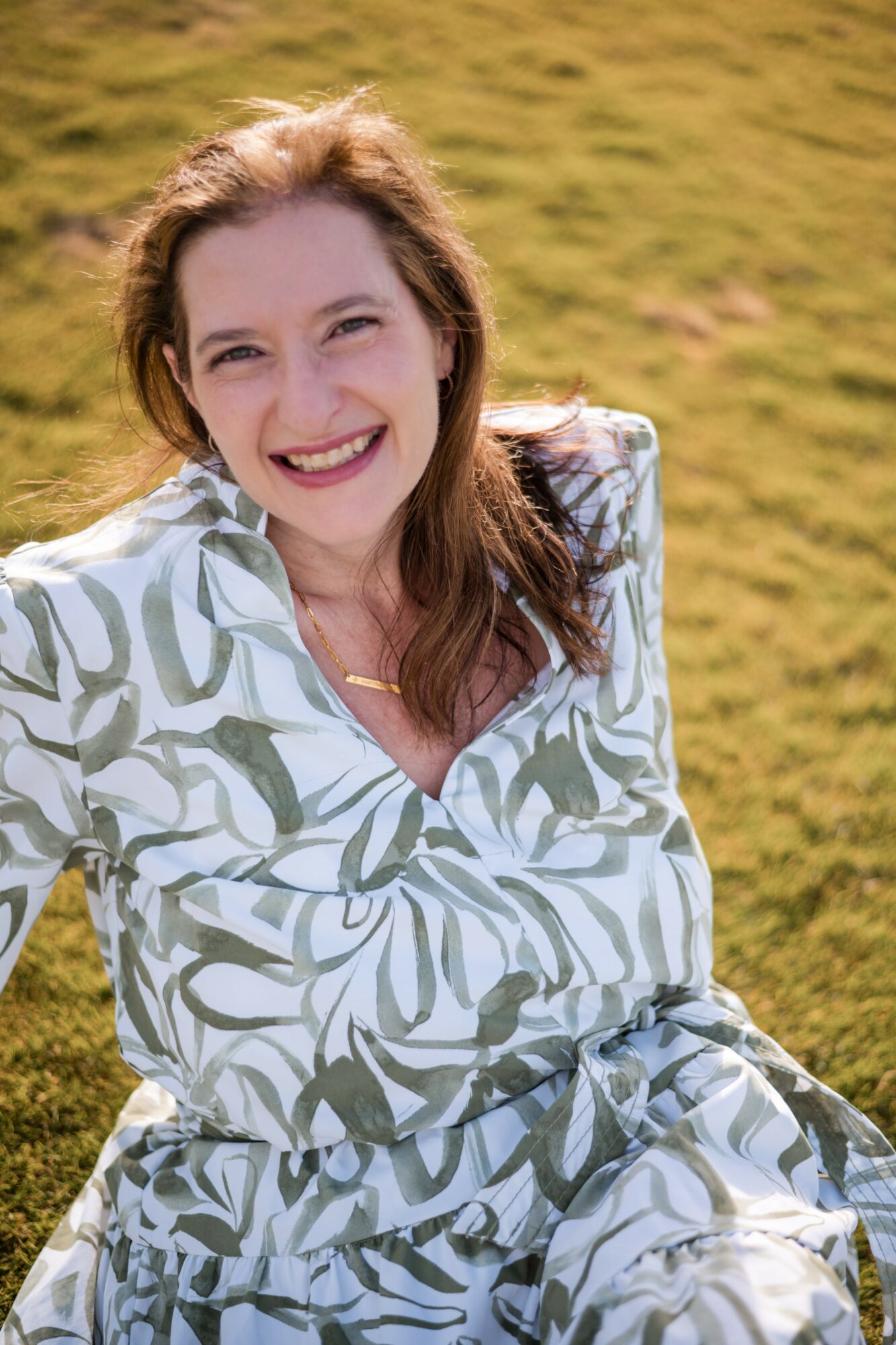 Smiling woman with long hair outdoors on grassy field, wearing a patterned dress, sunlight on face.