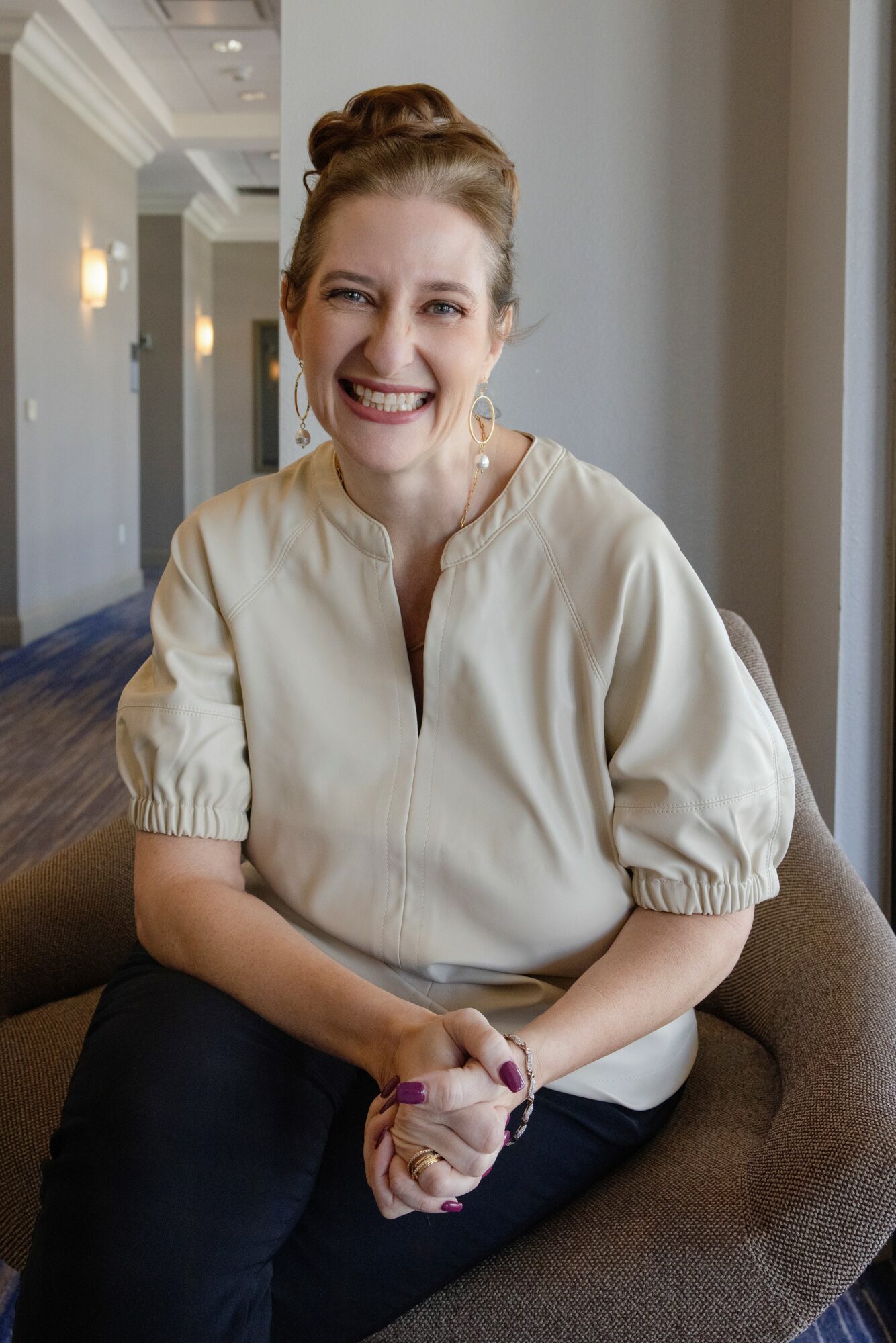 Smiling woman with hair in a bun, wearing a light-colored blouse, sitting on a chair indoors near a window.