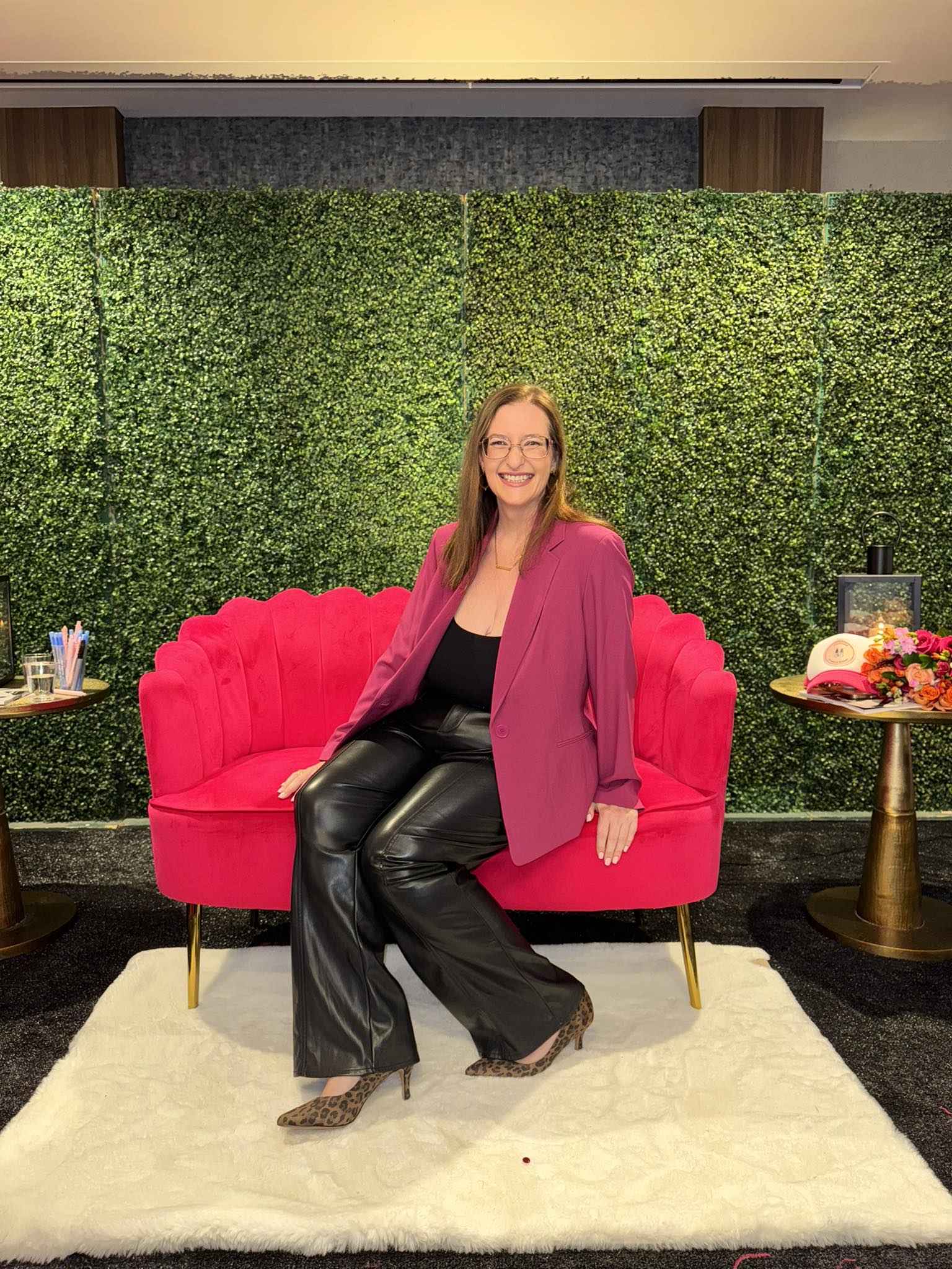 Woman sitting on a pink sofa with a green hedge background, wearing a pink blazer and black pants, smiling.