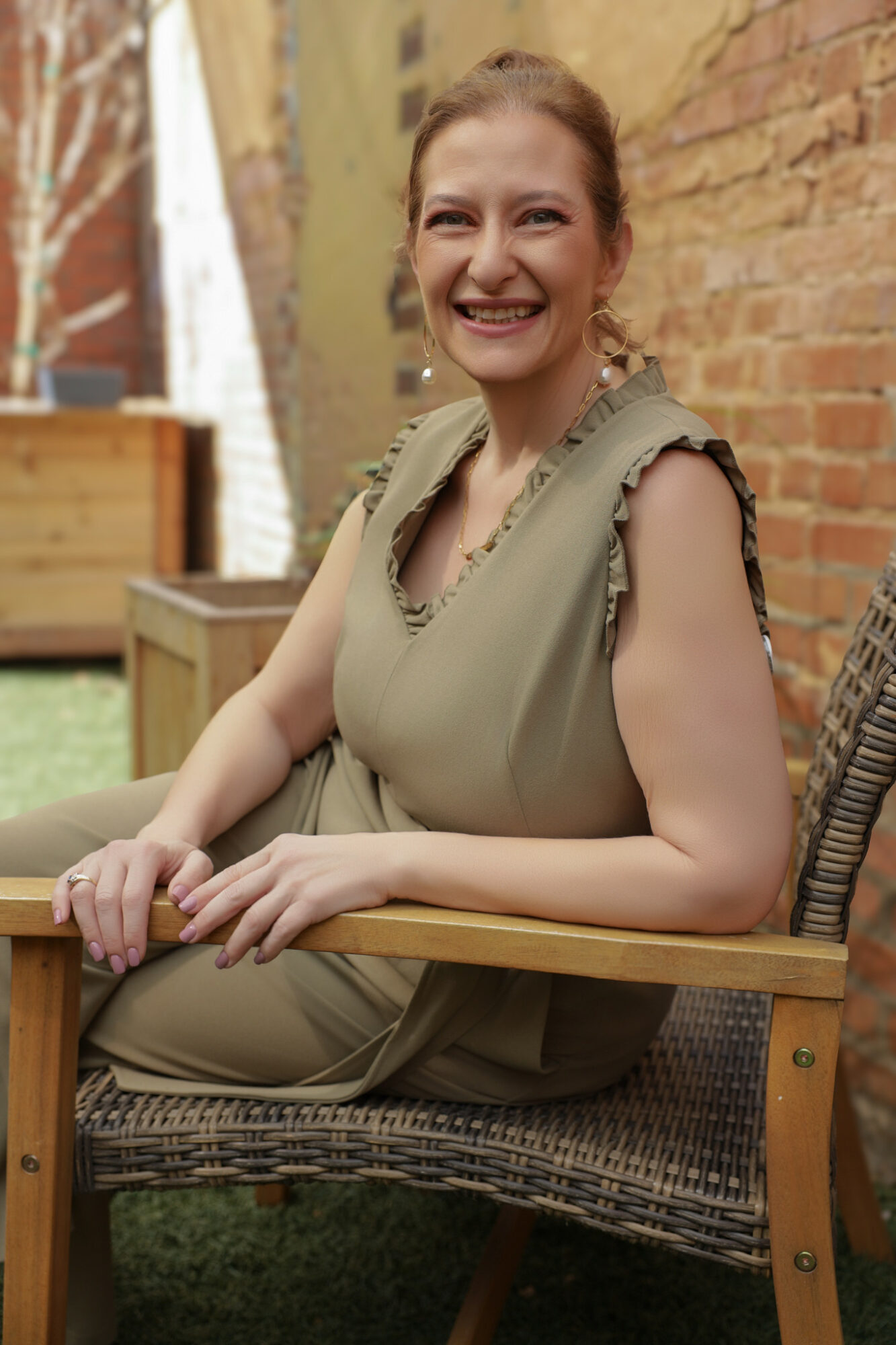 Smiling woman sitting outdoors at a wooden table, with a brick wall and greenery in the background.