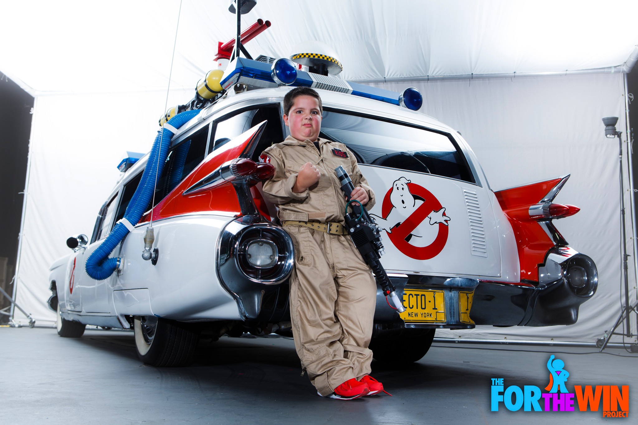 Person in Ghostbusters costume standing in front of a Ghostbusters-themed vehicle inside a studio, holding a prop weapon.